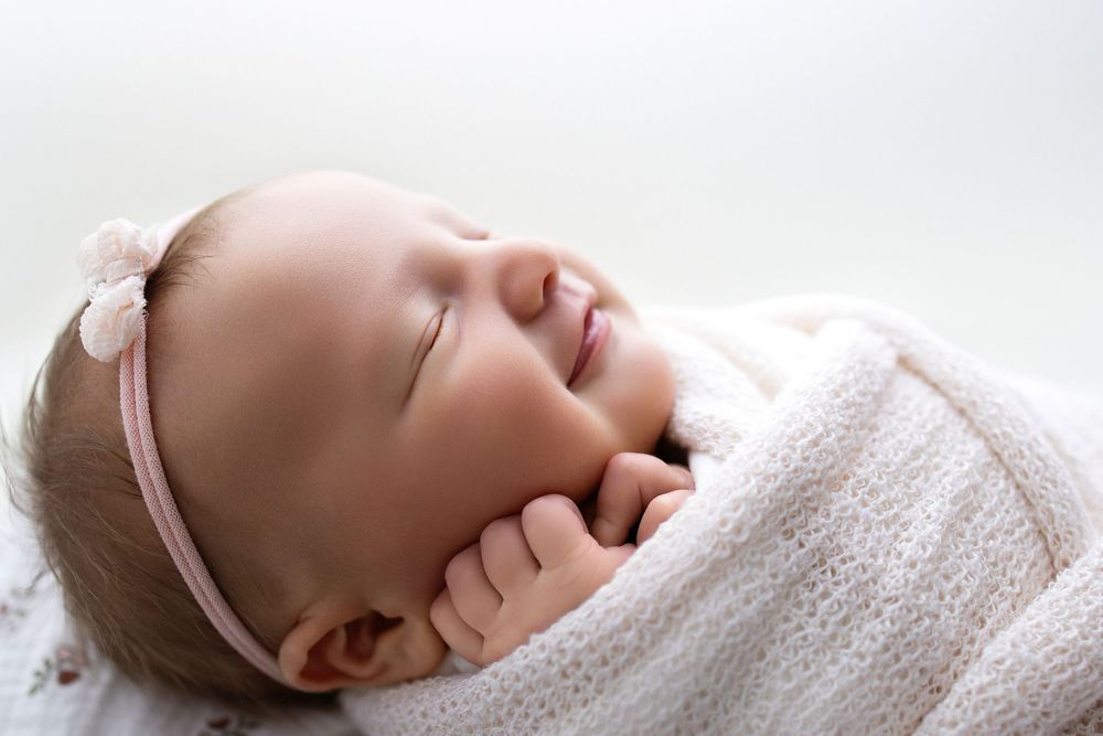 Peaceful sleeping baby wrapped in soft white blanket wearing pink headband smiles during naptime.