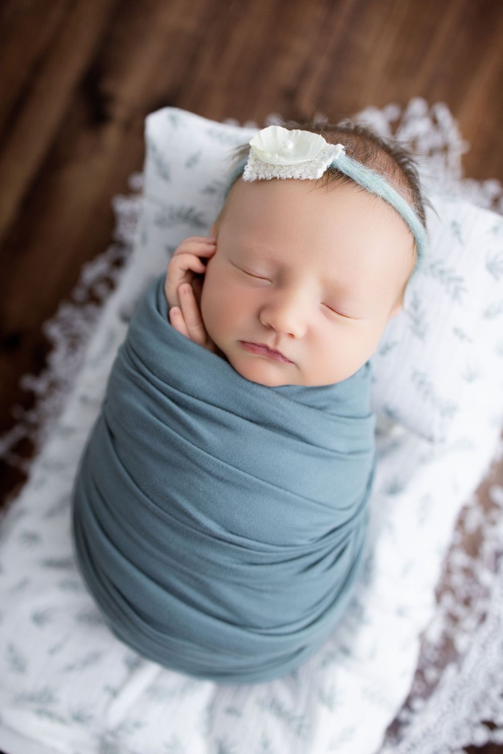 Newborn baby wrapped in soft dusty blue fabric swaddle sleeping peacefully on white textured backdrop.