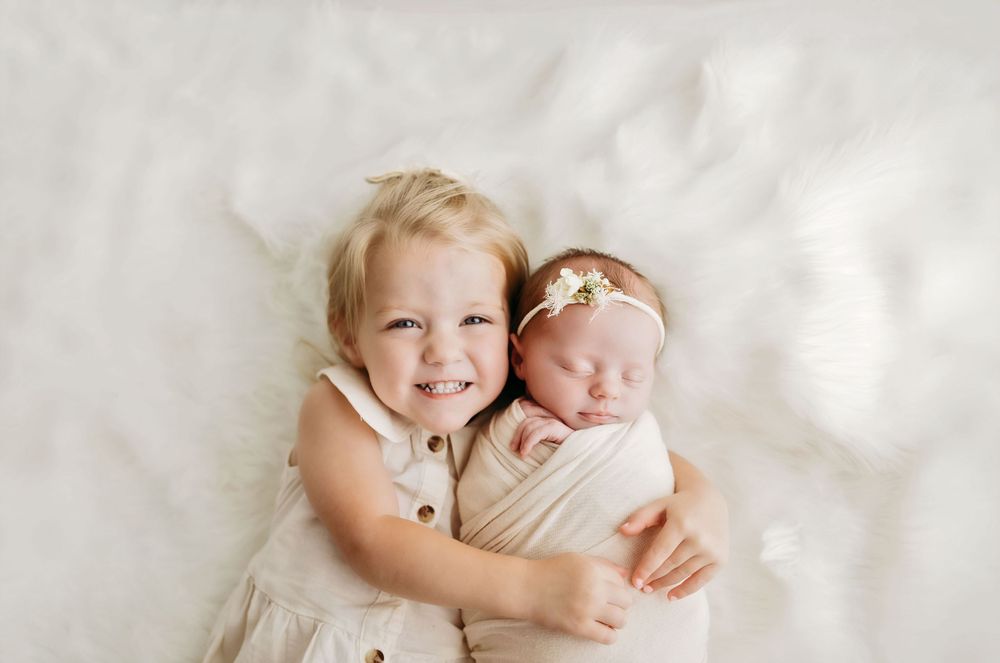 Two siblings in white outfits lying on white fabric, one hugging the other while smiling during a portrait session.