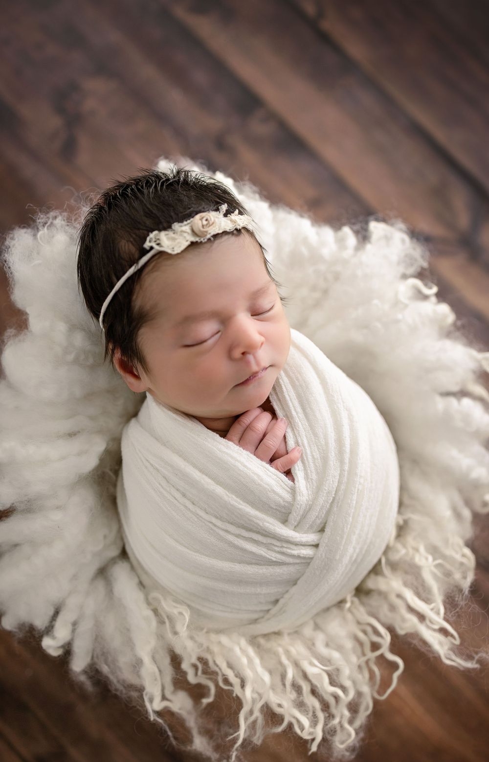Peaceful sleeping newborn wrapped in soft white fabric laying on fluffy white blanket against wooden floor background.