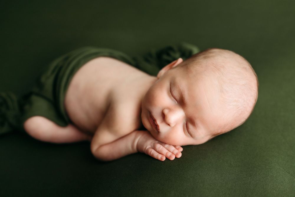 Peaceful sleeping newborn baby curled up on soft green fabric in classic newborn photography pose.