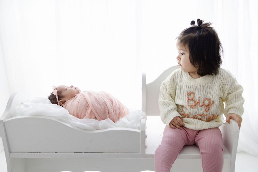 Two children in pink and cream outfits, with one sitting on a white bed while the other rests beside them.