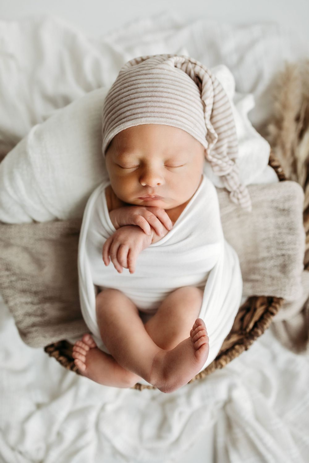 Sleeping newborn wrapped in white swaddle wearing striped beanie lies peacefully on soft textured bedding.