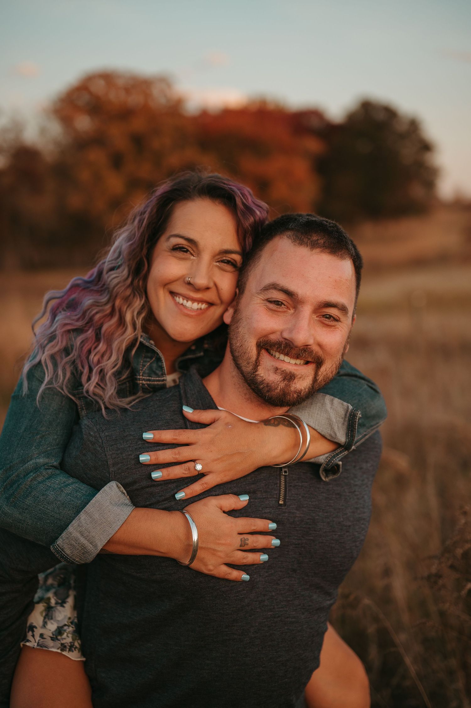 Couple shares joyful moment in field during golden hour with autumn colors in background.