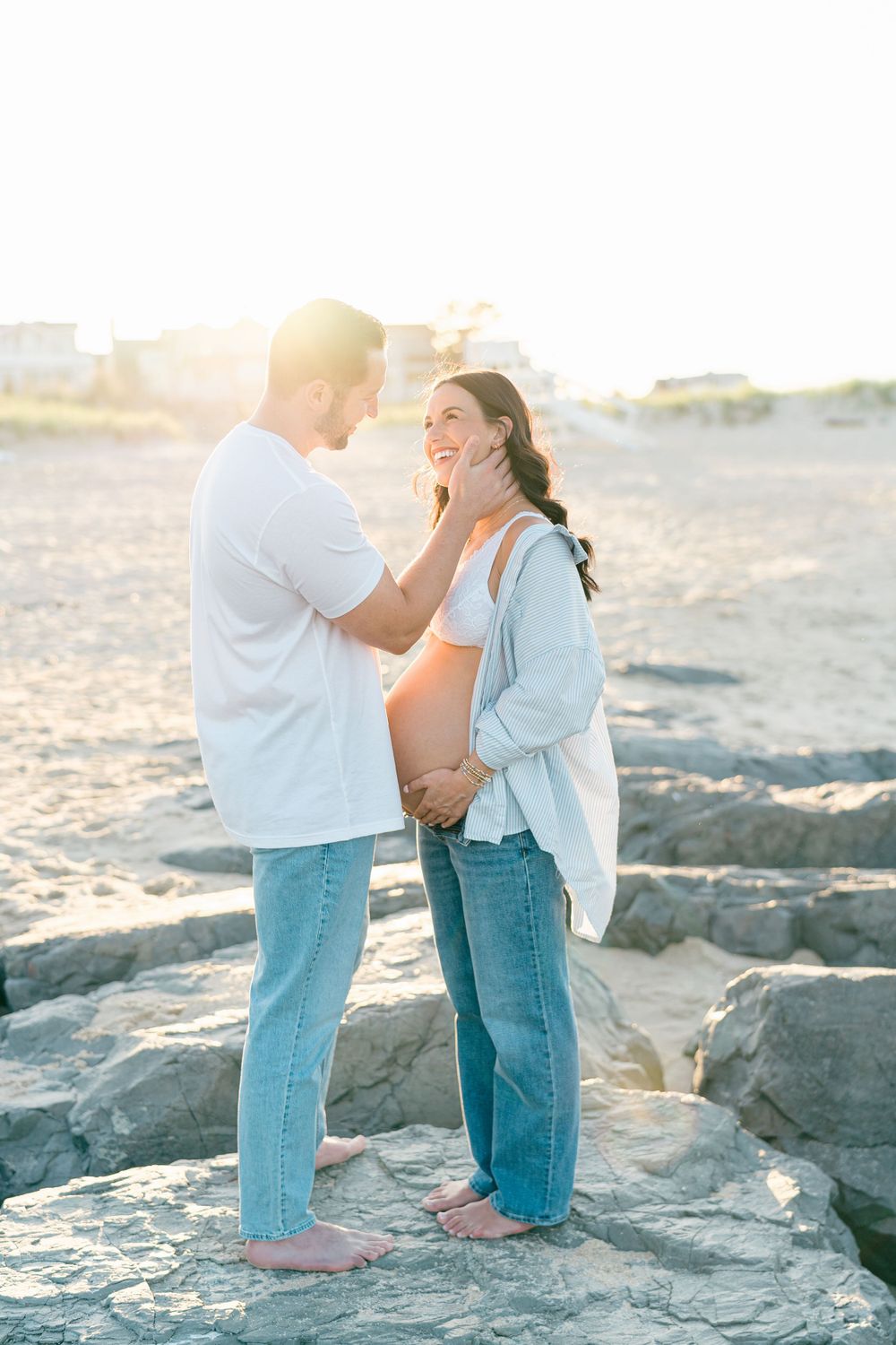 A man in jeans and a white shirt touches his pregnant wife's face while looking into her eyes on the beach.