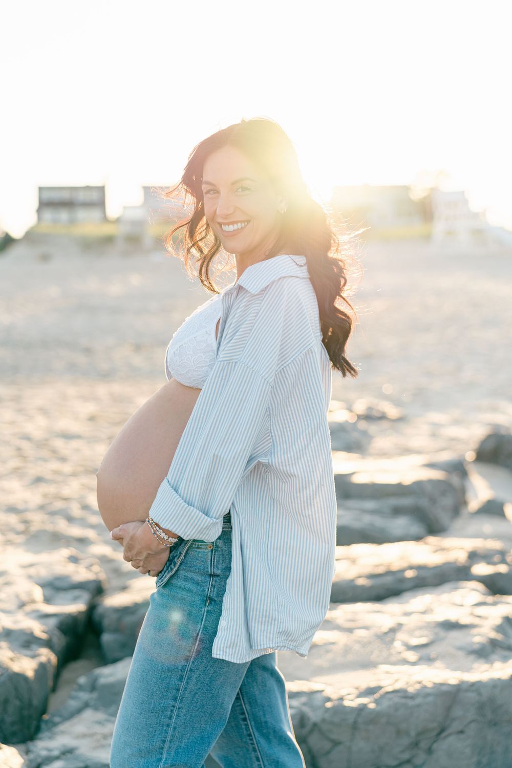 Pregnant woman standing on the beach, hands resting on her belly, with the sunset sky as her backdrop in Monmouth County, NJ.