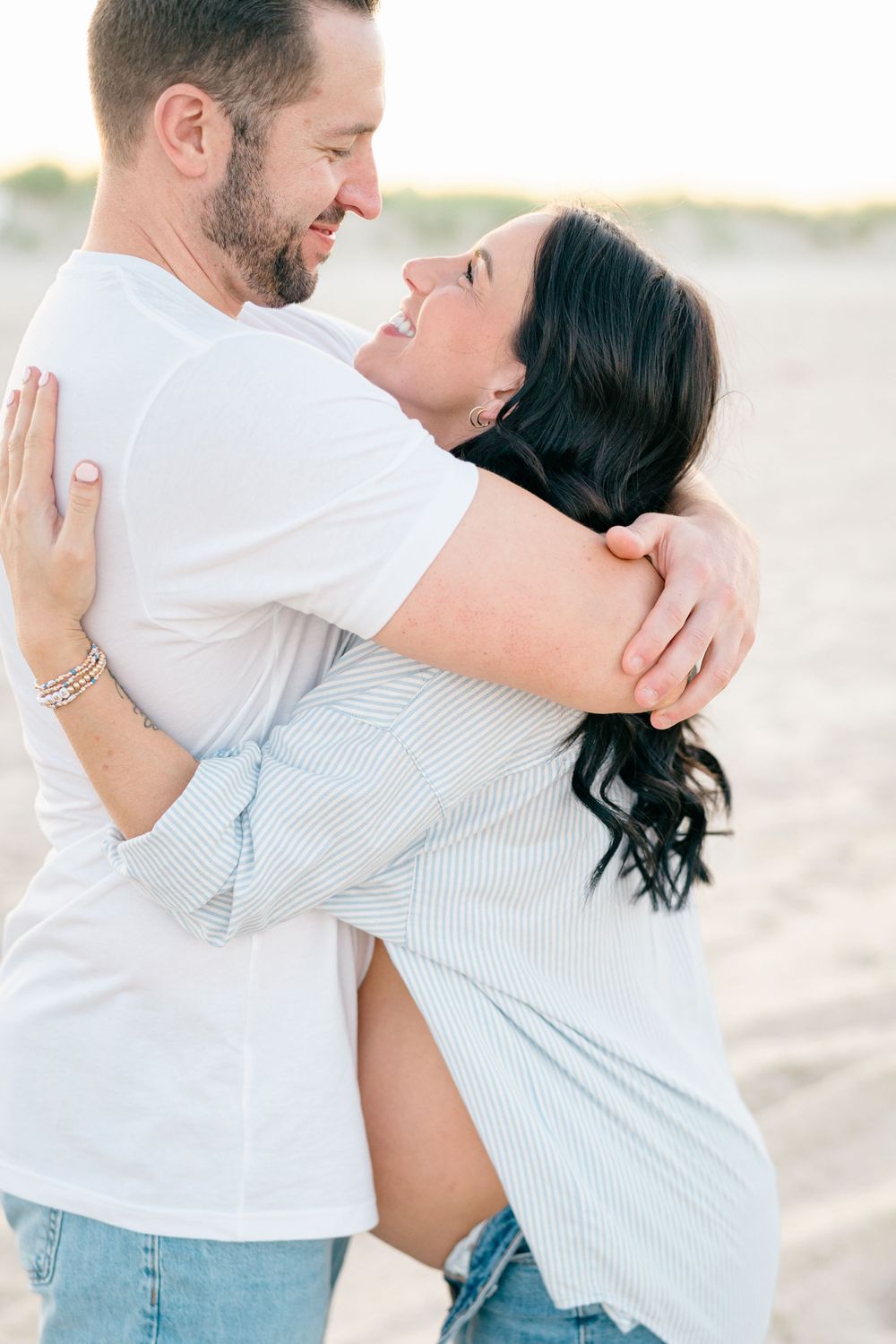 A couple hugs on the beach in spring lake NJ with sandy dunes behind them.