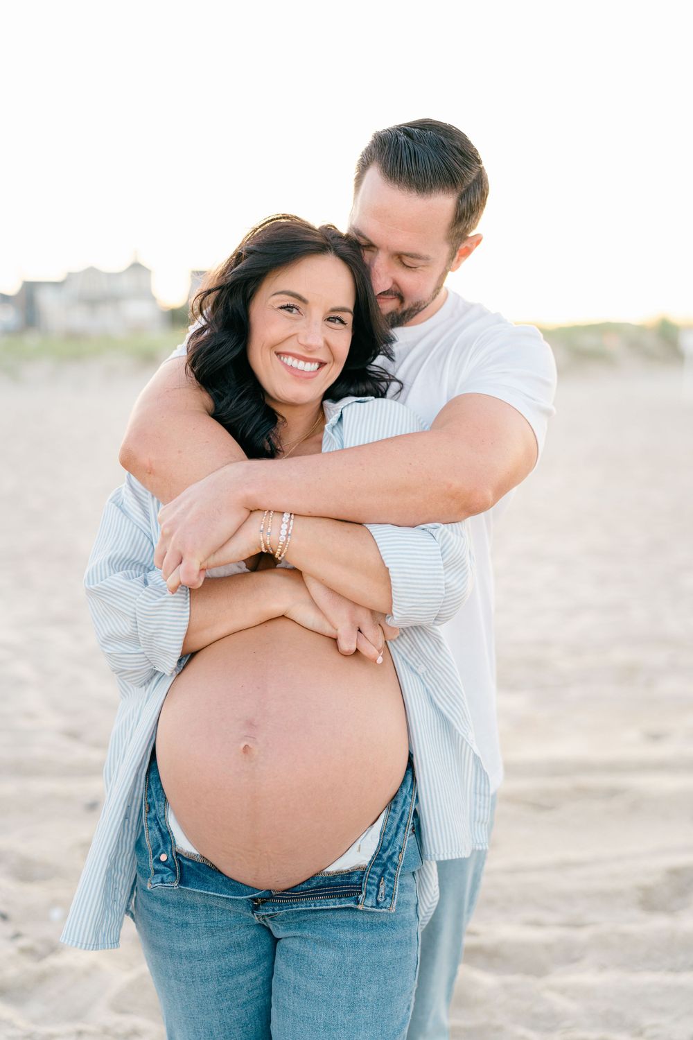 A man hugs a woman from behind on the sandy beach.