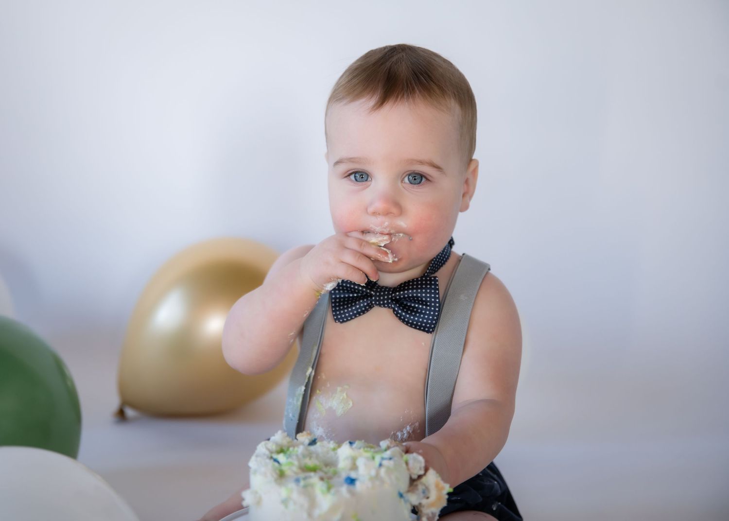 A toddler in suspenders and bow tie sits with party decor and balloons while enjoying birthday cake.