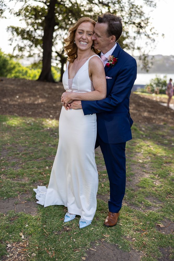 A joyful outdoor wedding scene of a couple embracing, with the bride in white and groom in navy blue suit.