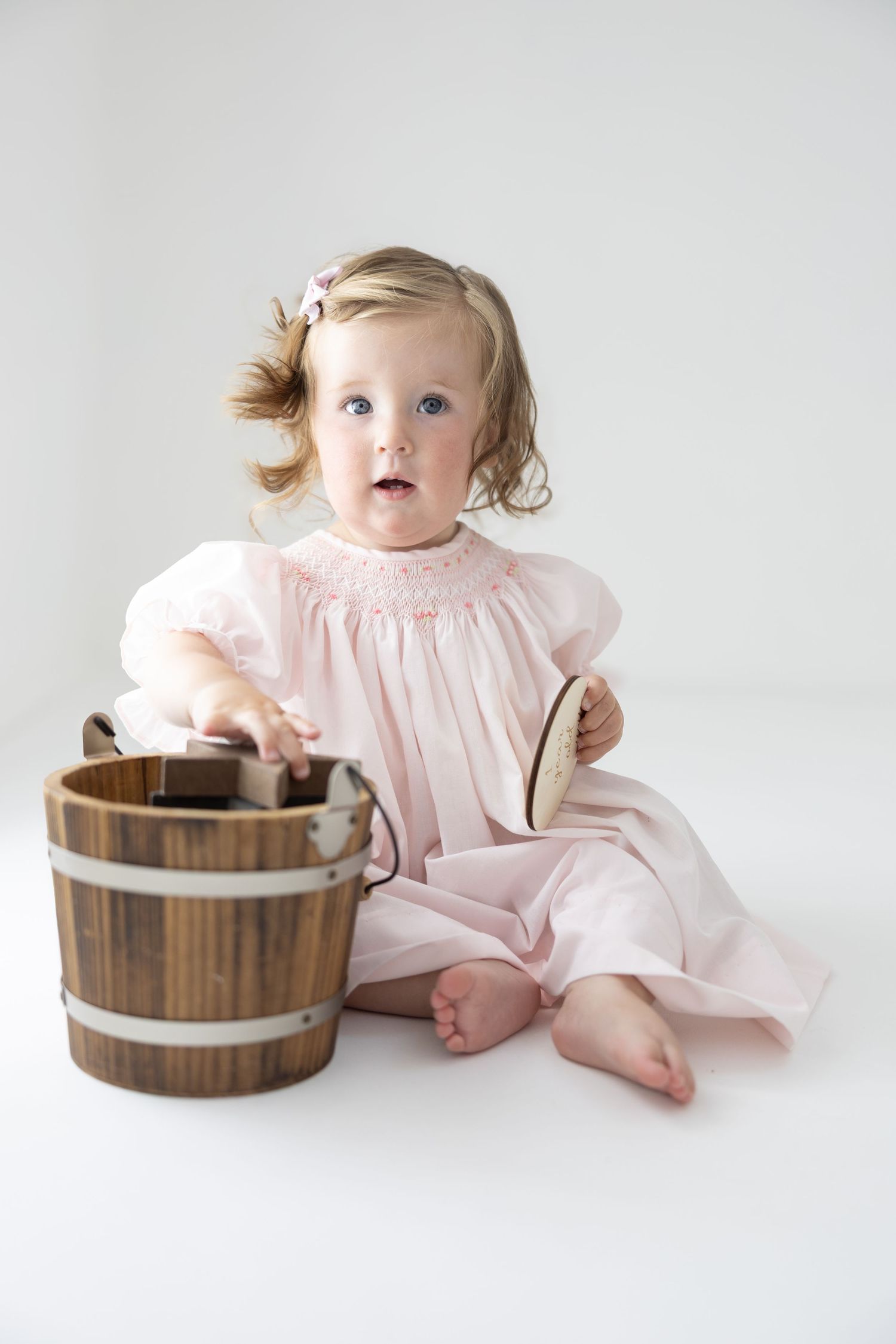 Toddler girl in pink smocked dress sitting with wooden bucket on white background.