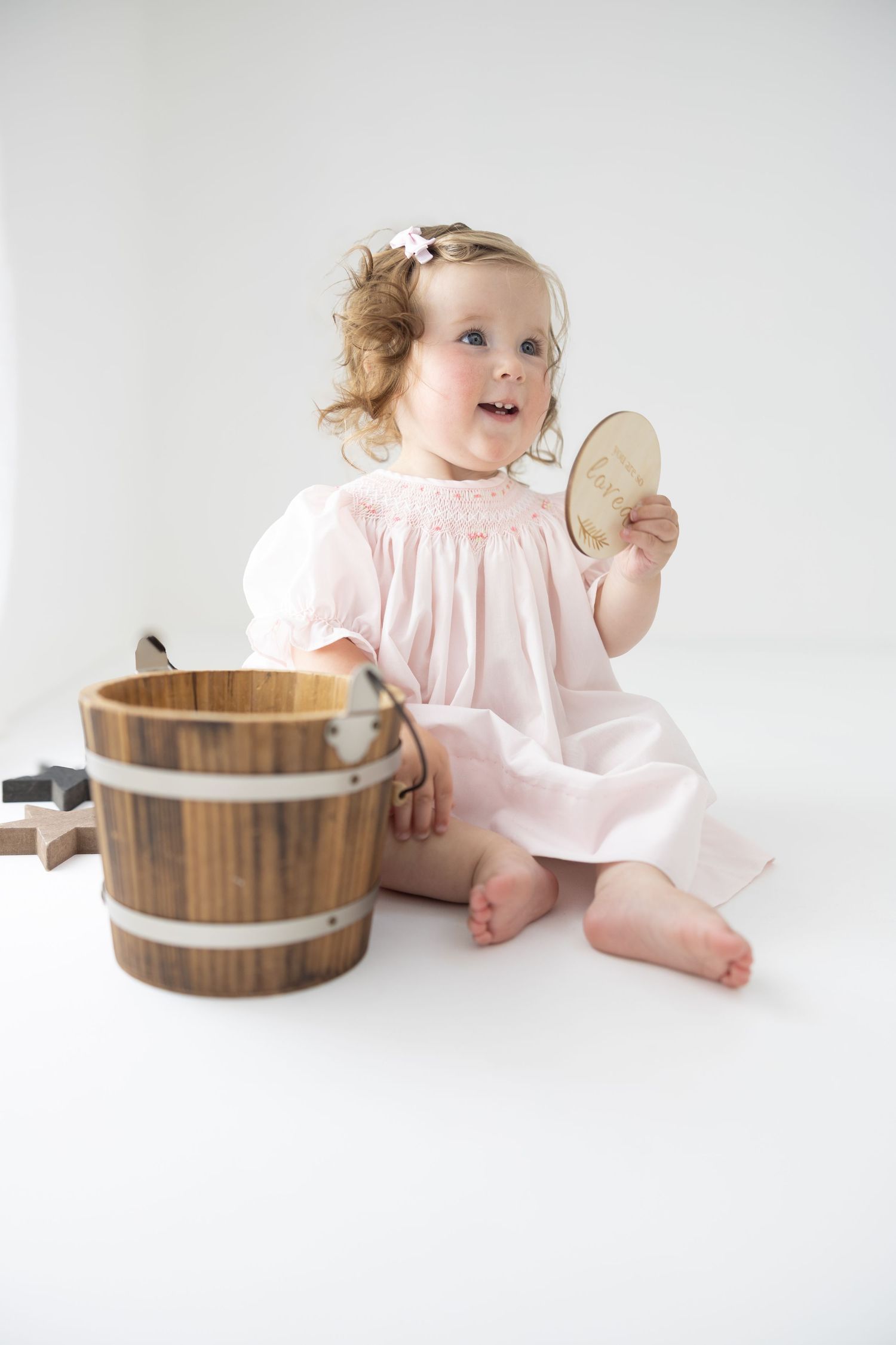 Toddler girl in pink smocked dress sitting on white floor holding wooden disc next to rustic wooden bucket.