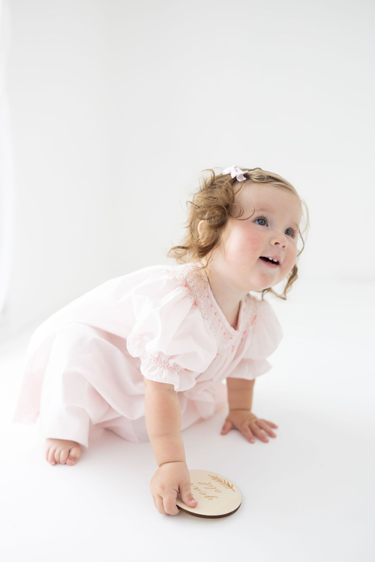 A baby girl in a pink dress crawls on a white floor, looking up curiously, with a wooden disc in hand.