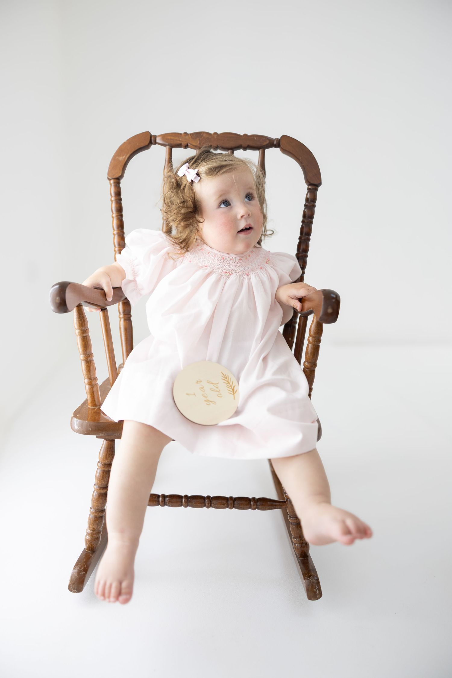 Toddler girl in pink smocked dress sitting in antique wooden rocking chair against white background.