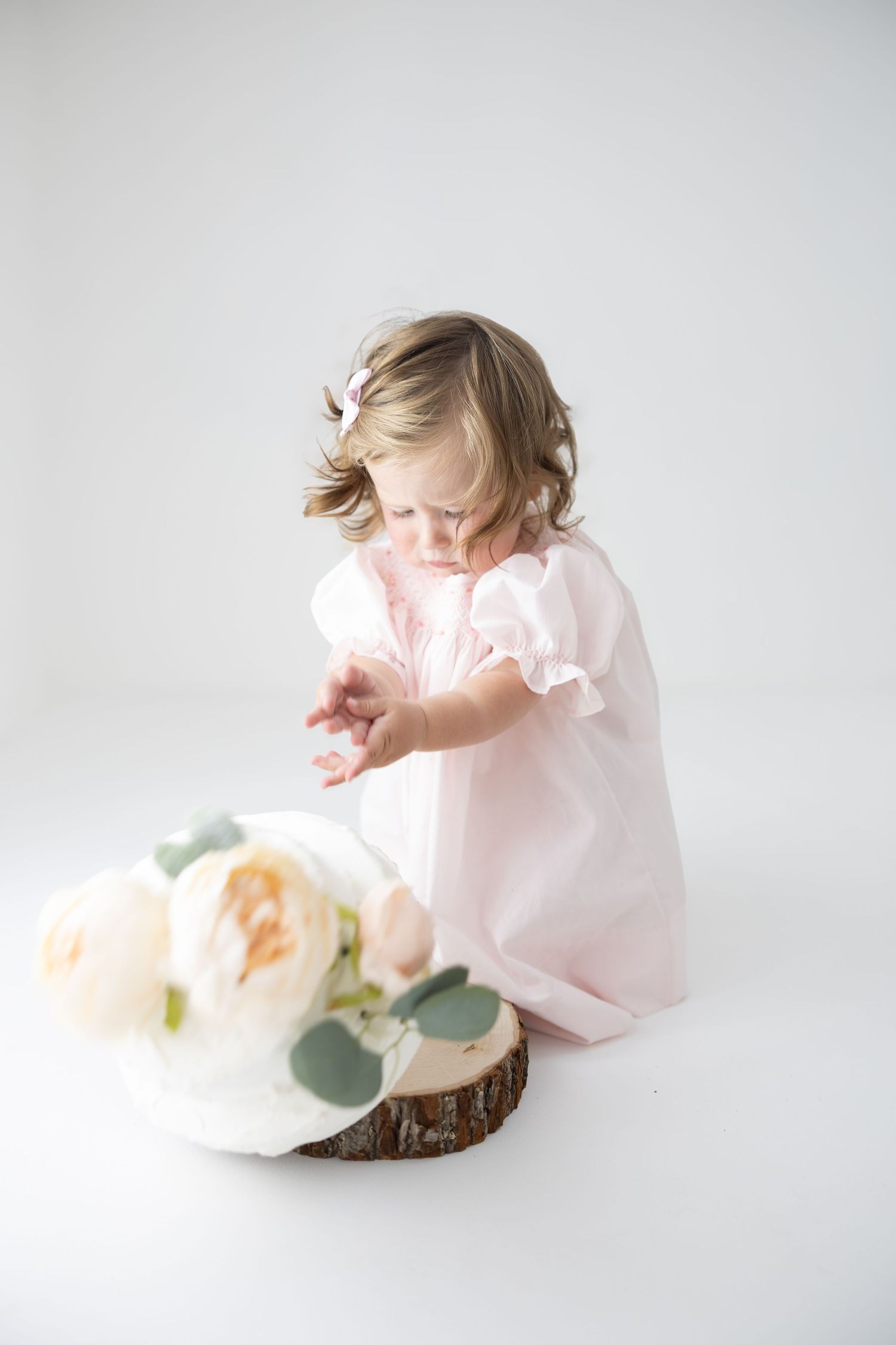 Toddler girl in pink dress reaching toward white roses on wood slice against white background.