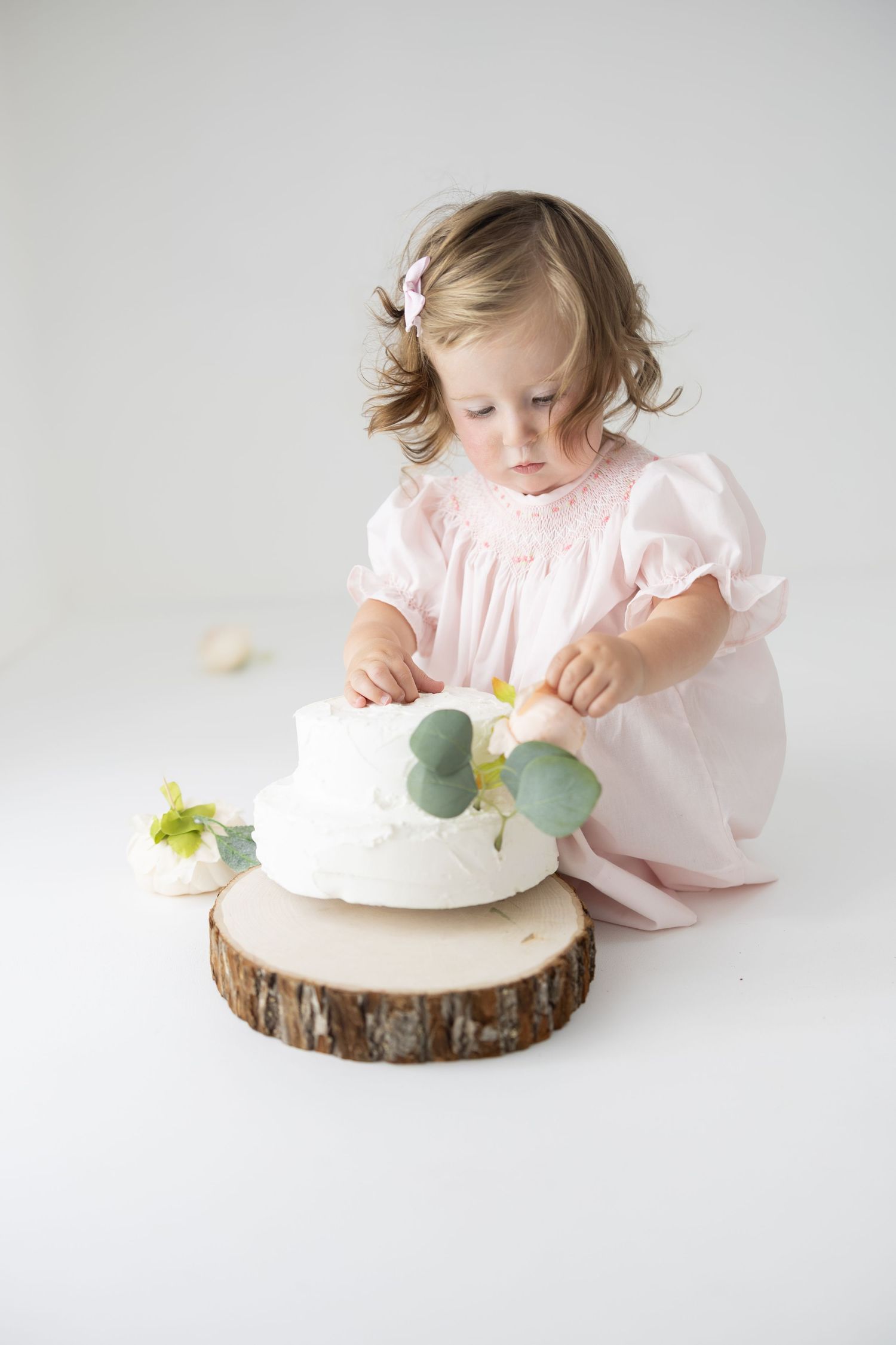 Toddler girl in pink dress touching a white two-tier cake with eucalyptus on a wood slice stand.