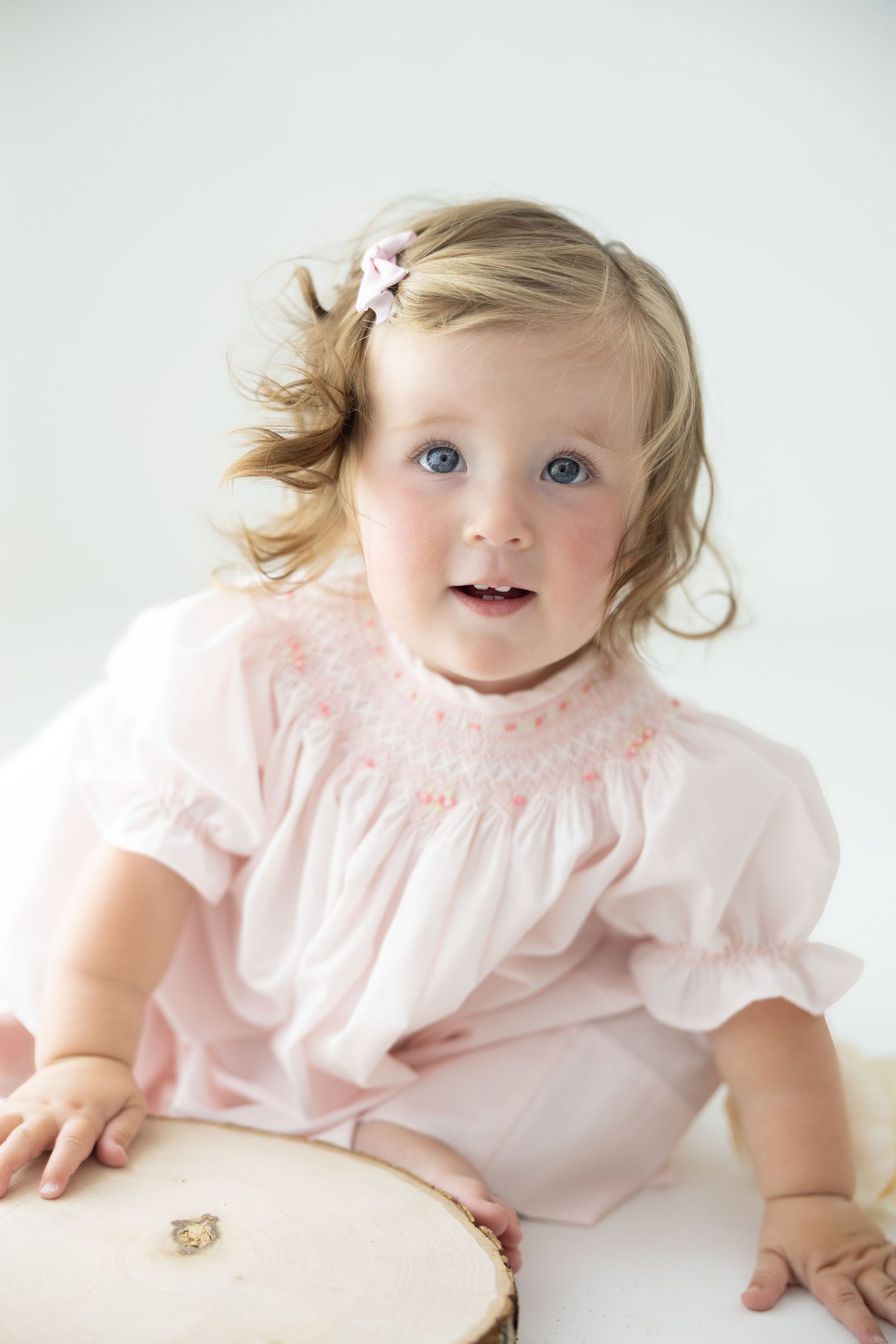 Adorable toddler girl with blue eyes in pink smocked dress and hair bow sits on white background.
