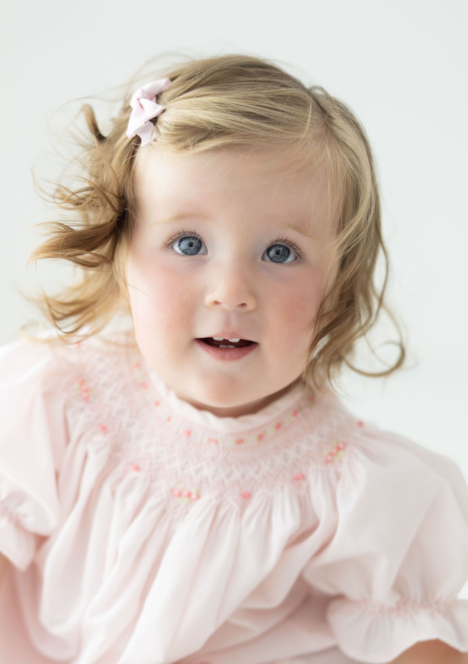 Adorable toddler girl with blue eyes and blonde hair wearing a pink smocked dress with a pink bow hair clip.