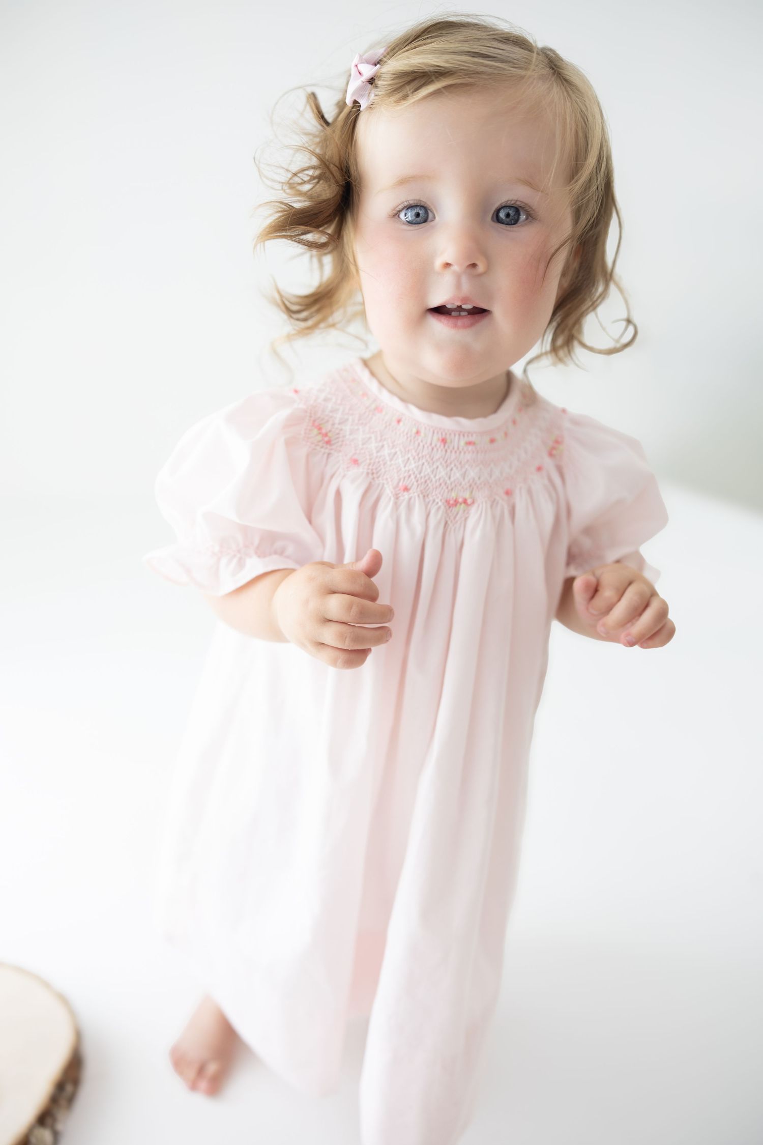 Toddler girl in pink smocked dress with puff sleeves and hair bow, standing against white background.