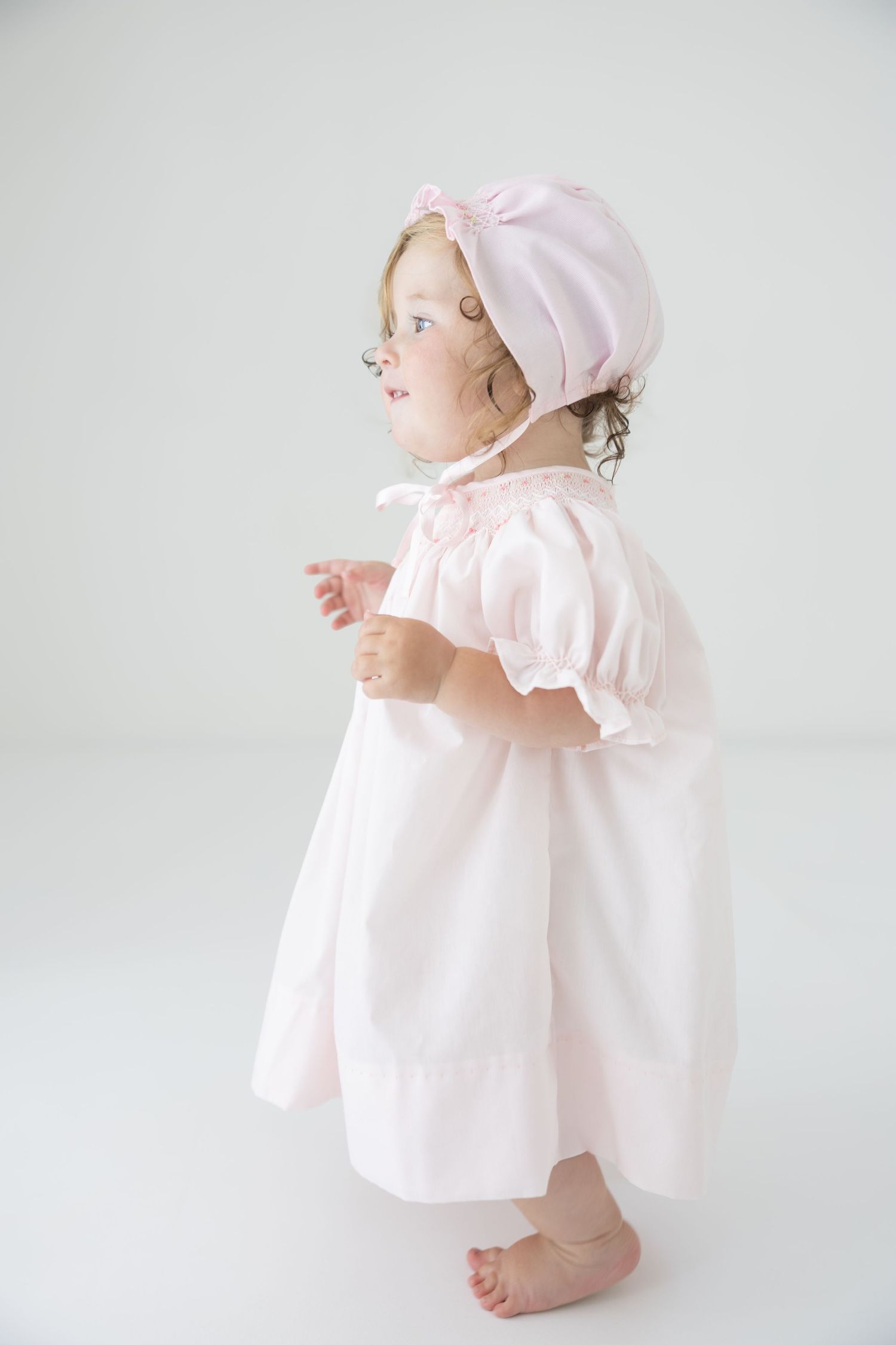 Toddler girl in pink dress and bonnet standing barefoot against white background.