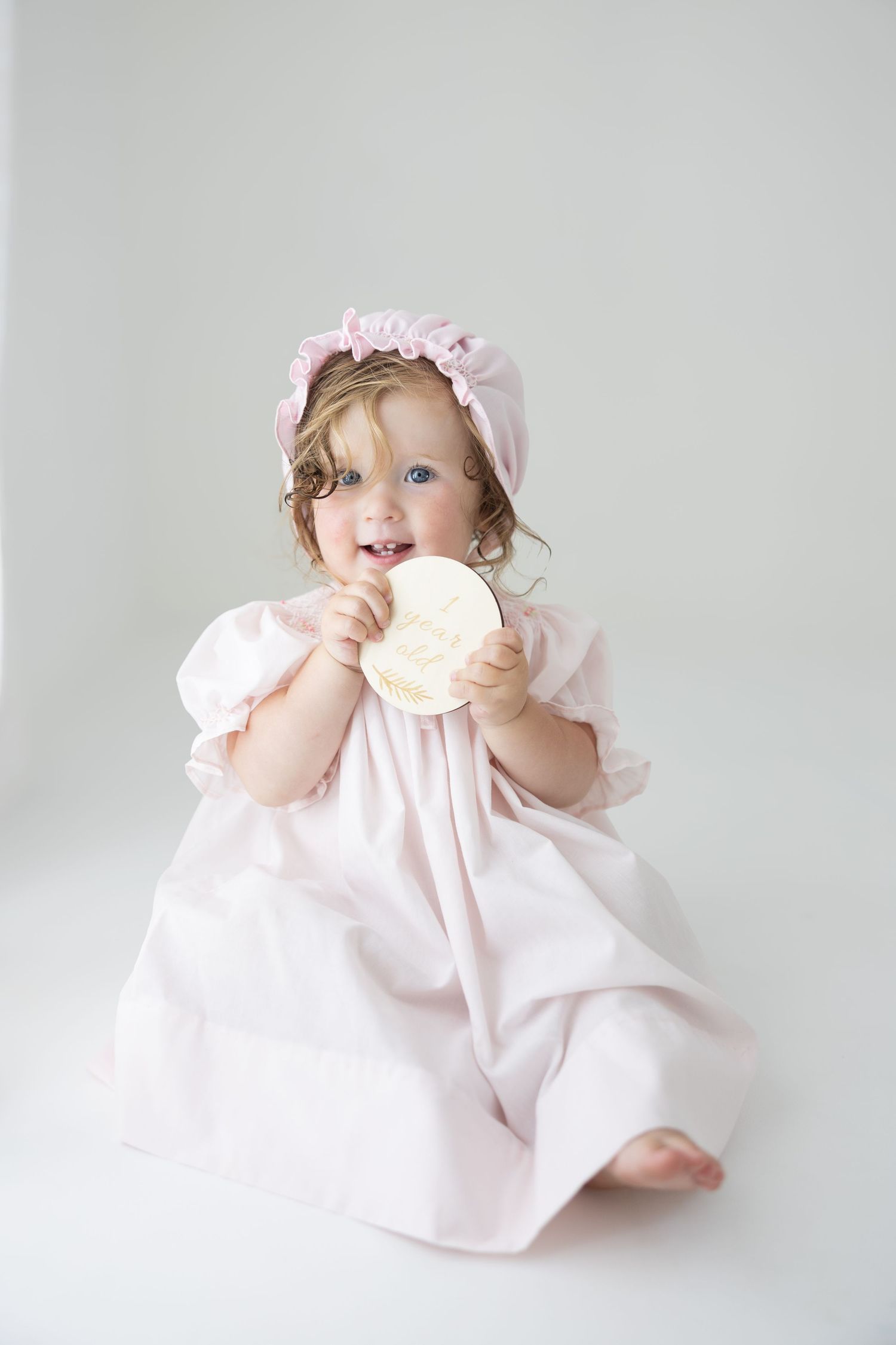 Toddler girl in pink dress and bonnet holding a large white chocolate coin, sitting on white background.