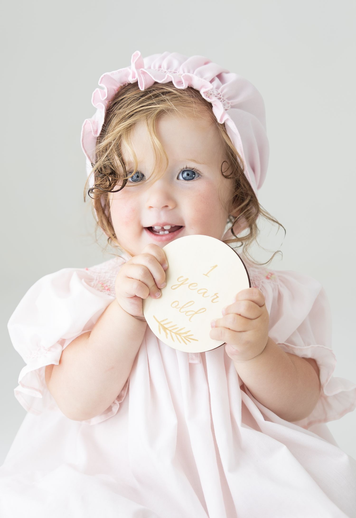 Adorable 1-year-old baby girl in pink dress and bonnet holding a wooden milestone disc, smiling with blue eyes.