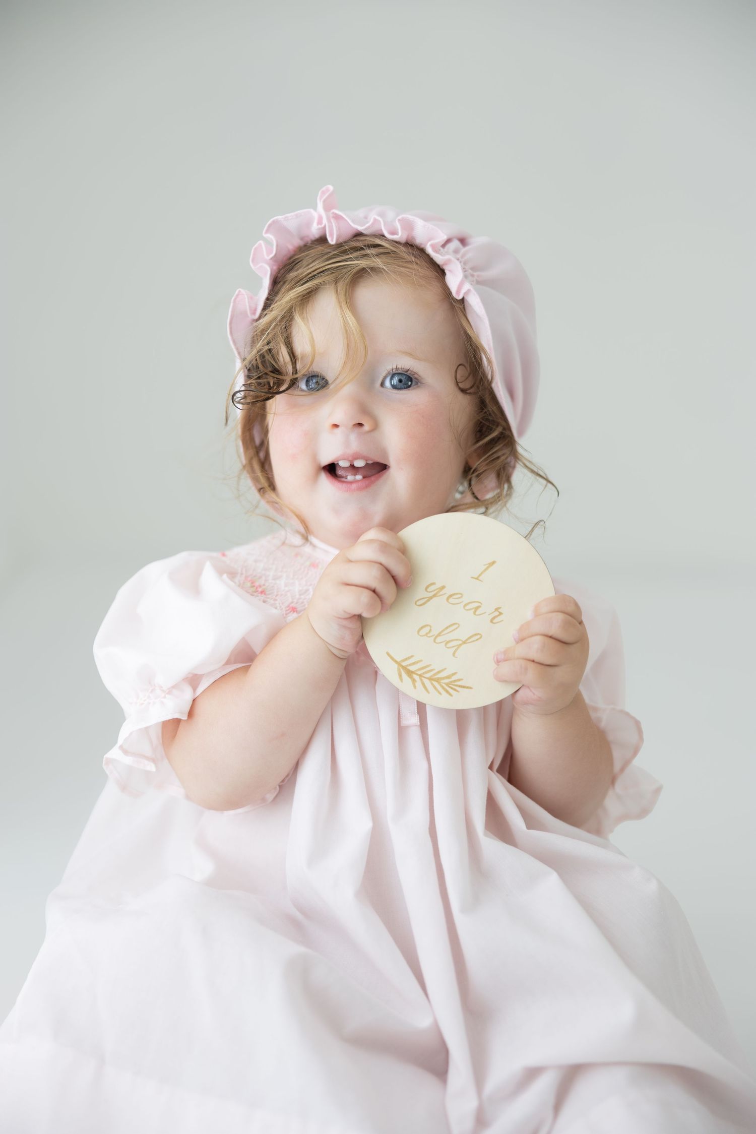 Smiling 1-year-old baby girl in pink dress and bonnet holding a wooden milestone disc.