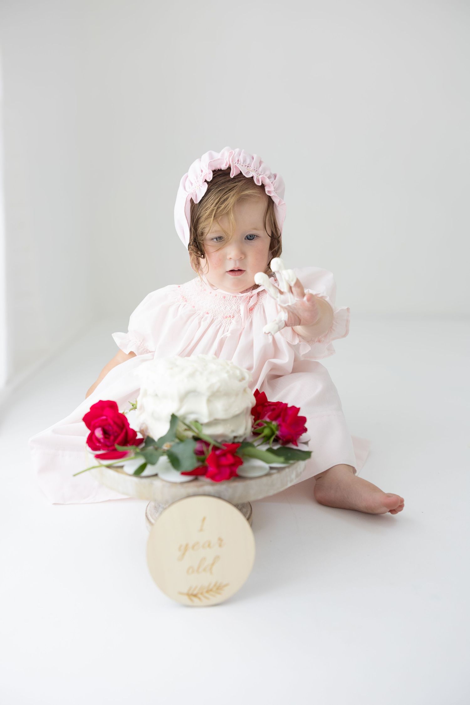 A baby girl in a pink dress and bonnet sits beside a white cake with red roses for her 1st birthday.
