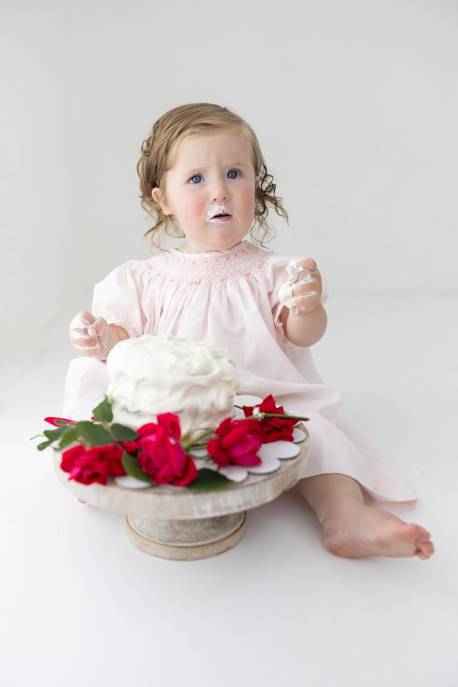 Baby girl in pink dress eating first birthday cake with white frosting and red roses on white background.