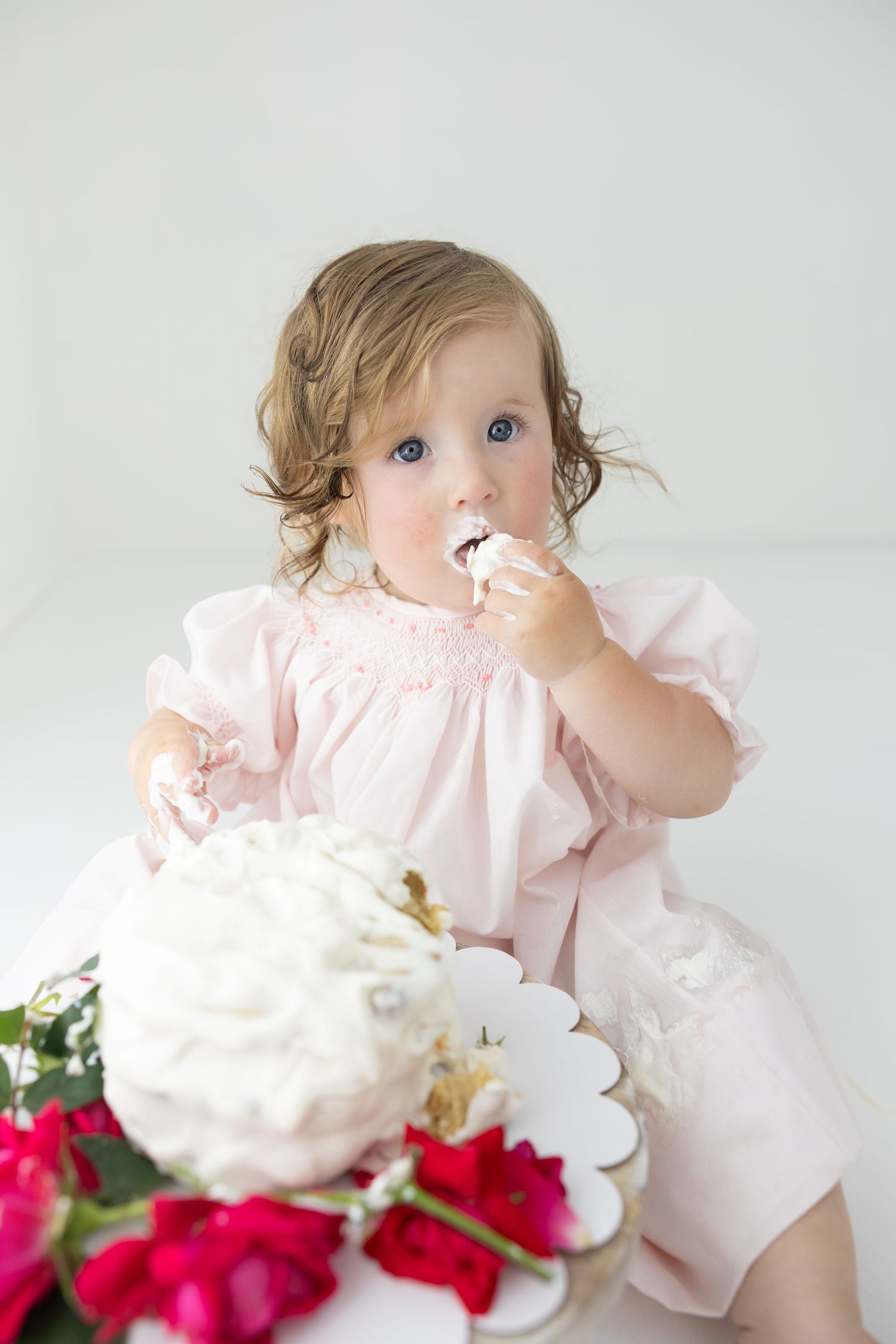 Toddler girl in pink smocked dress eating birthday cake with white frosting surrounded by red roses.
