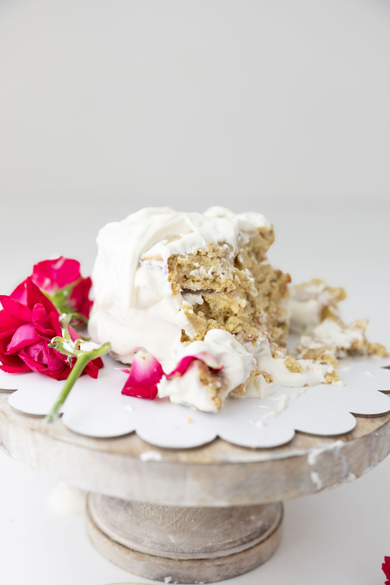Partially eaten white frosted cake on a rustic wooden cake stand with red roses beside it.