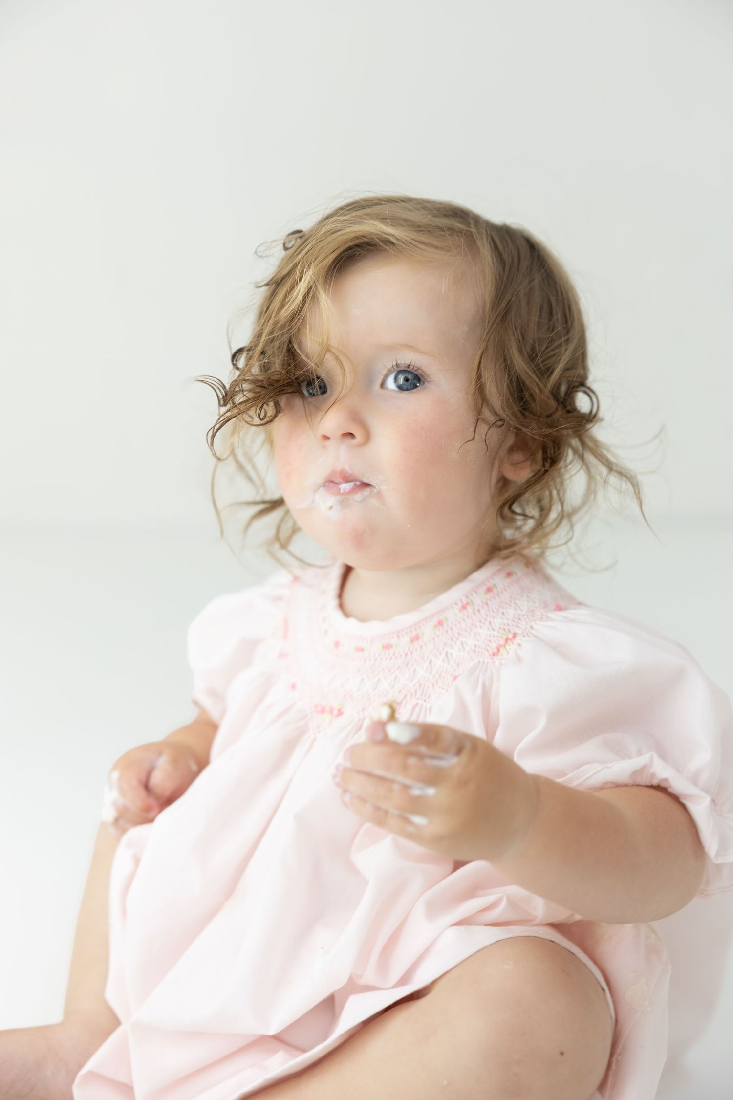 Toddler girl in pink smocked dress with curly hair and blue eyes, sitting with food on her face.