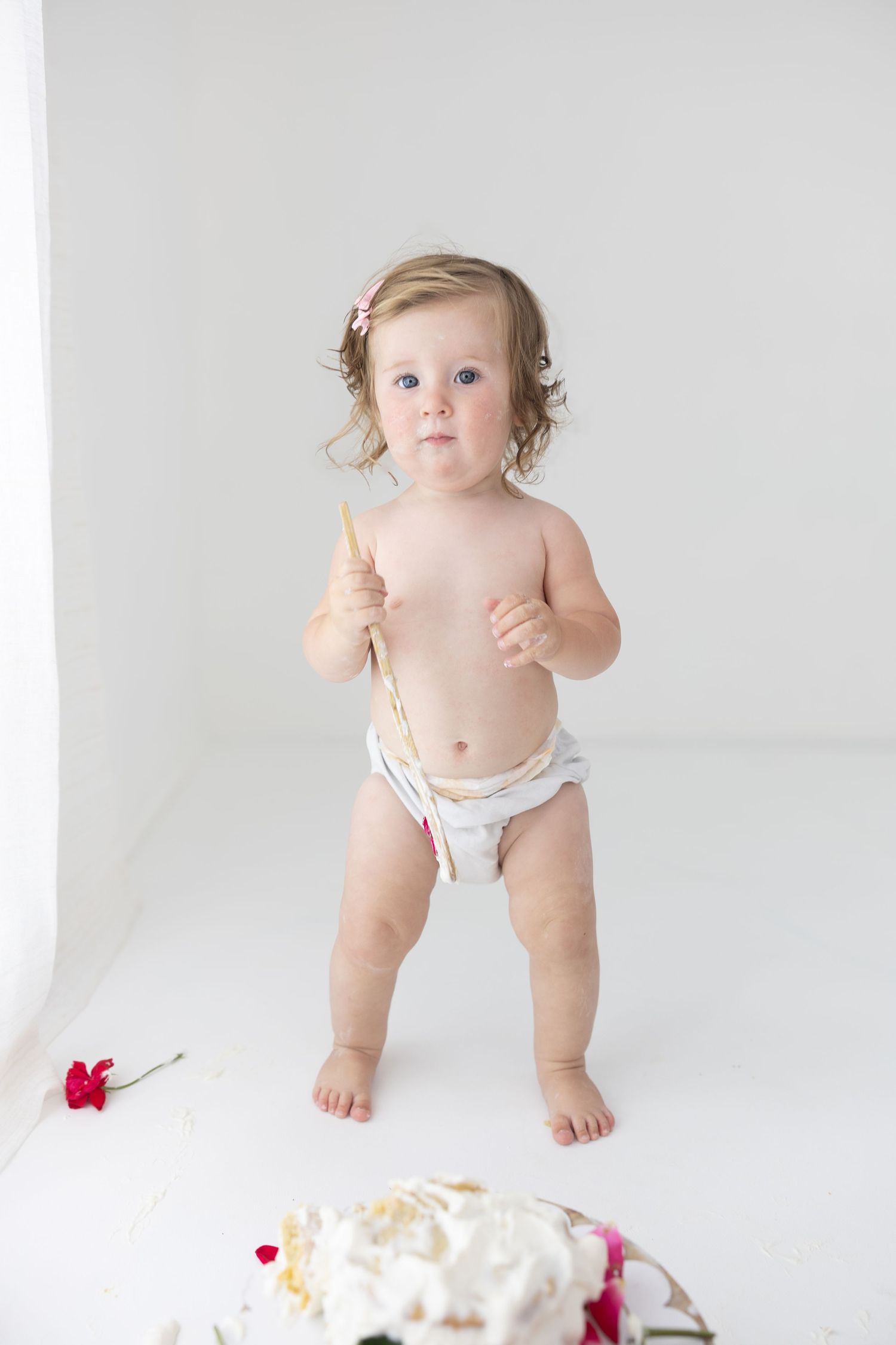 A baby girl in a diaper holds a gold spoon near a smashed first birthday cake on a white background.