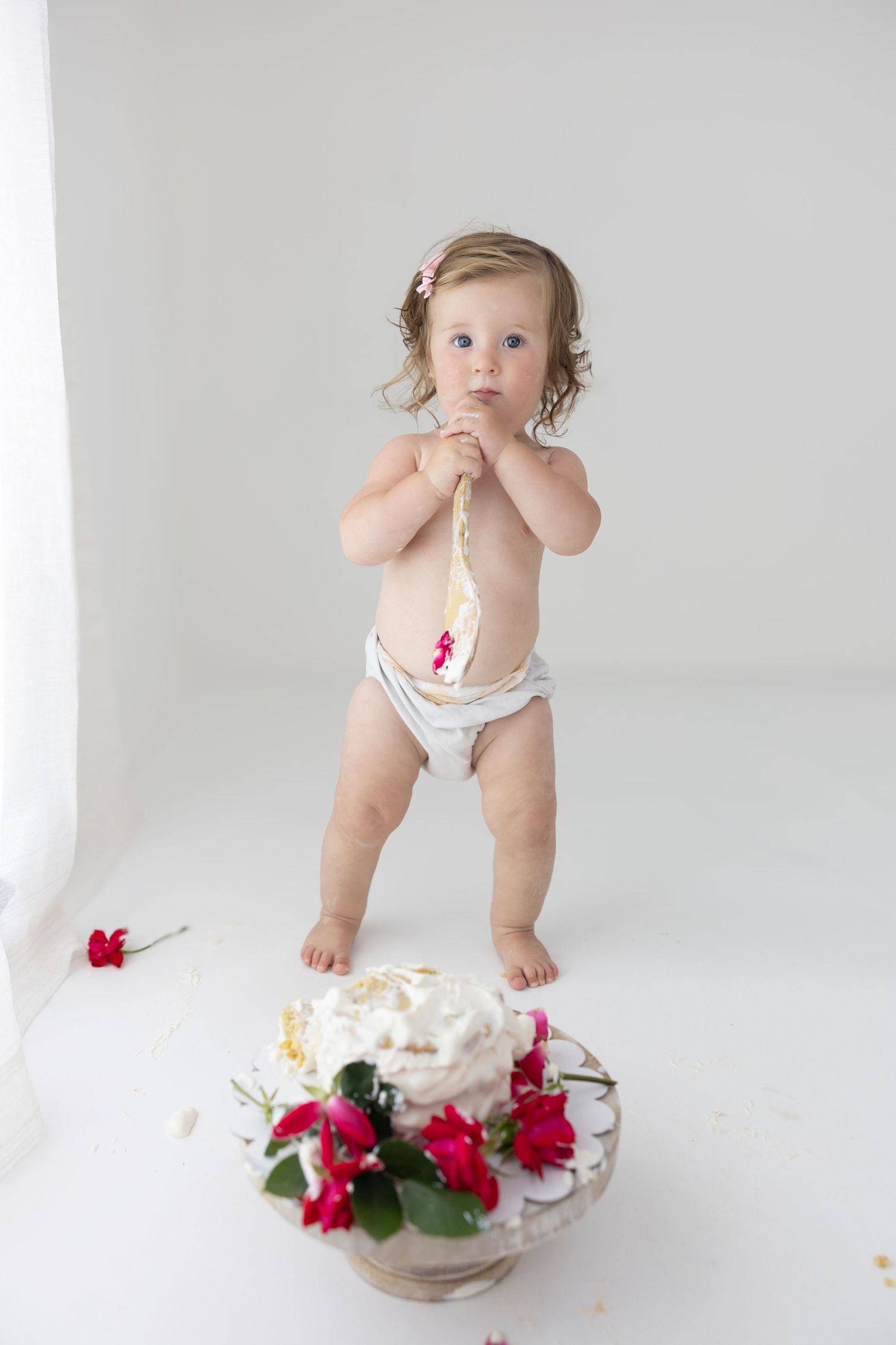 Toddler in diaper standing by a white cake with red roses during a first birthday cake smash photoshoot.