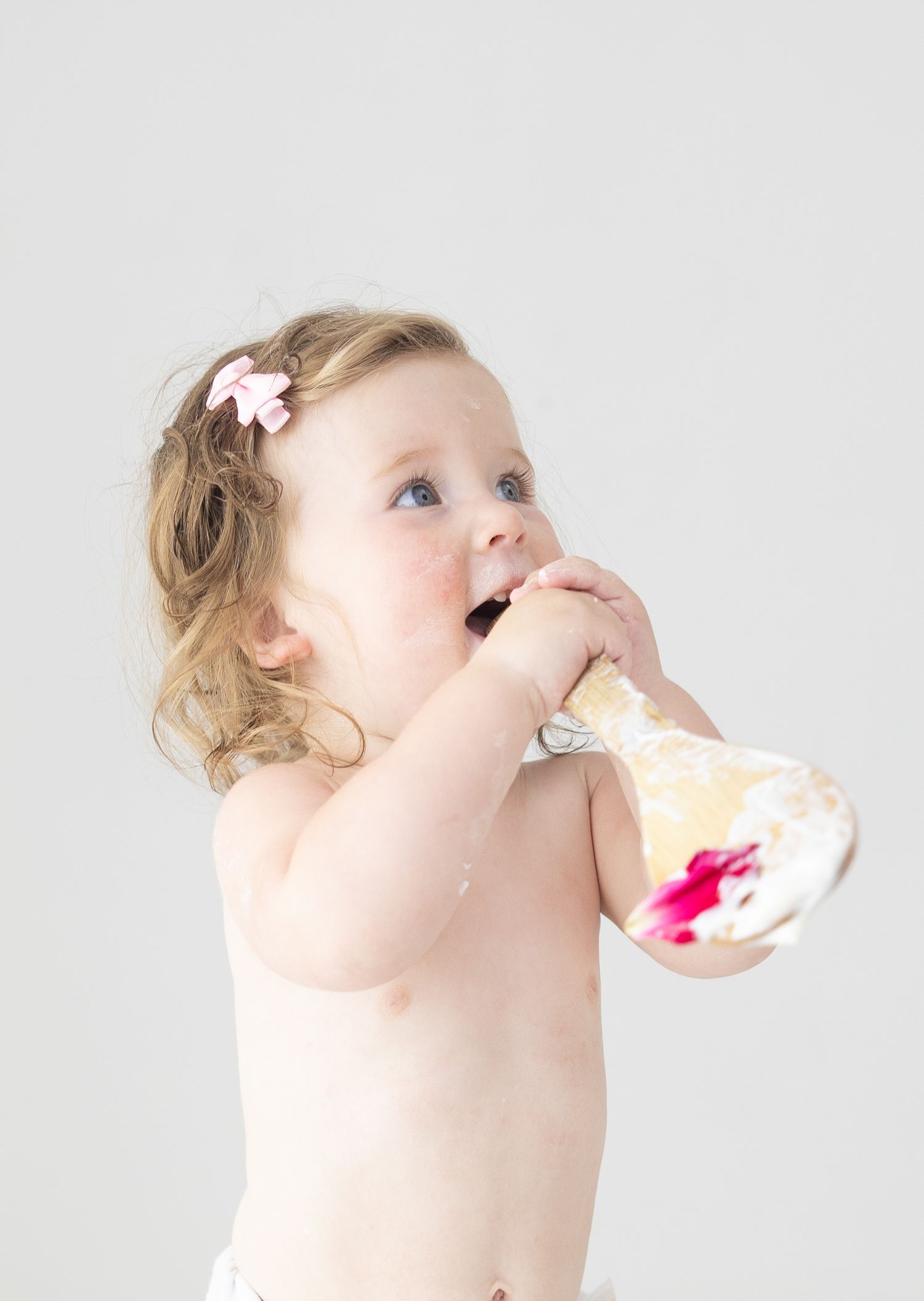 A cute baby girl with a pink bow eating cake from a glass dish during her first birthday smash cake photoshoot.