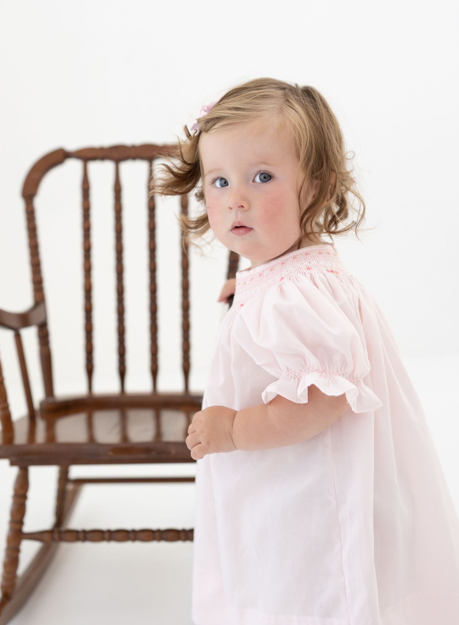 Toddler girl in pink smocked dress standing beside a wooden rocking chair, looking over her shoulder.