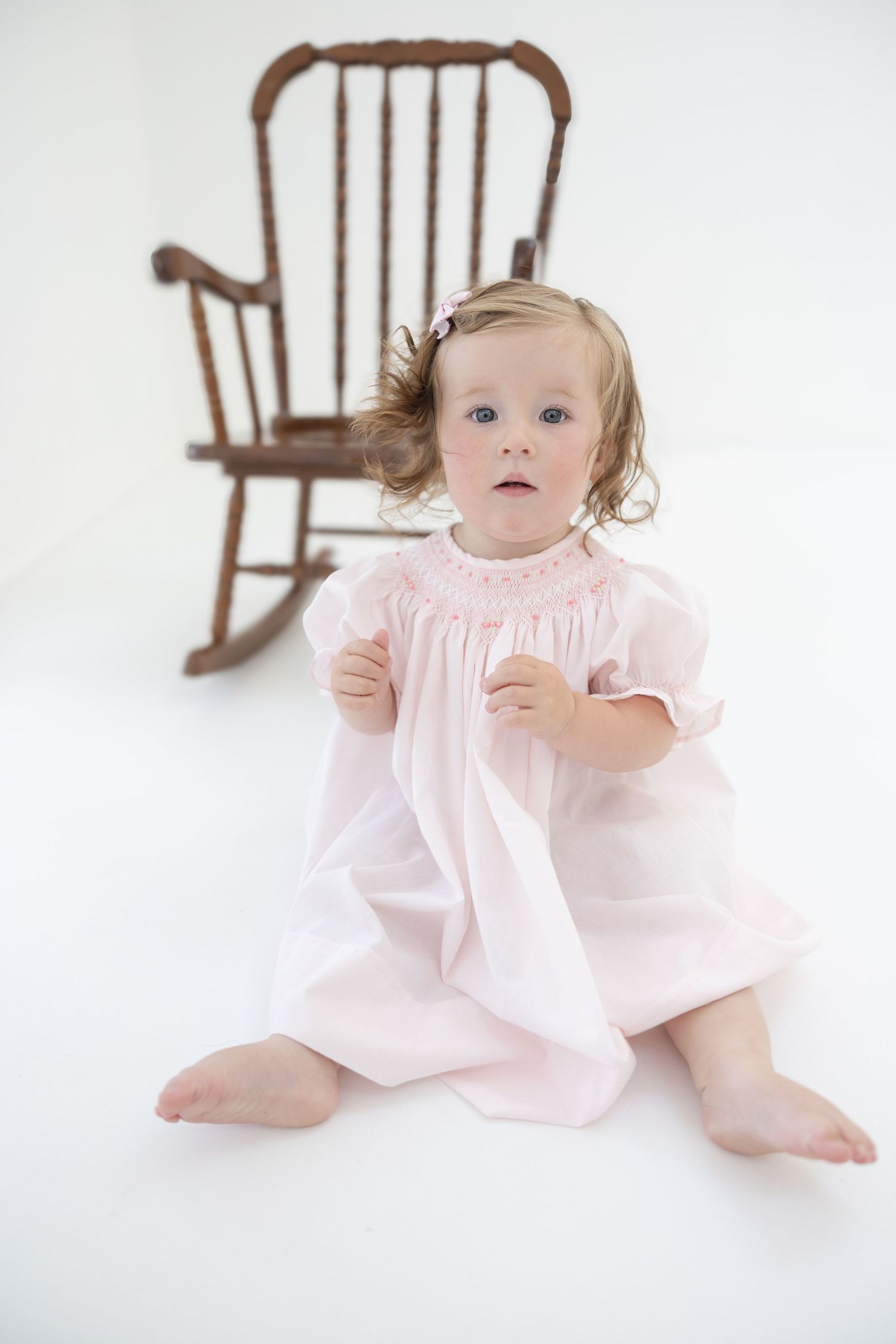 A toddler girl in a pink smocked dress sits barefoot on a white floor with a vintage wooden rocking chair behind her.