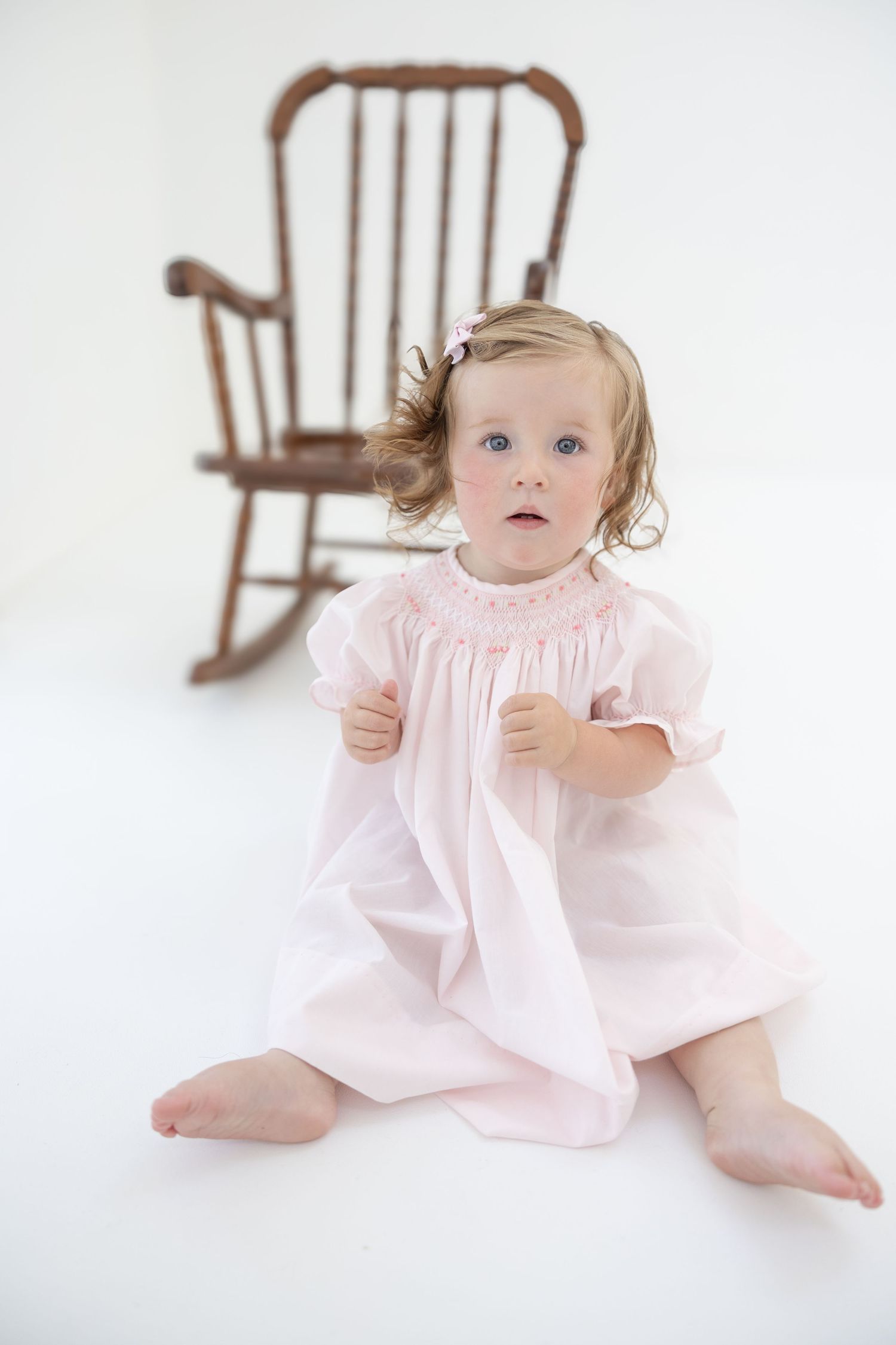 A toddler girl in a pink smocked dress sits barefoot on white floor with a wooden rocking chair behind her.