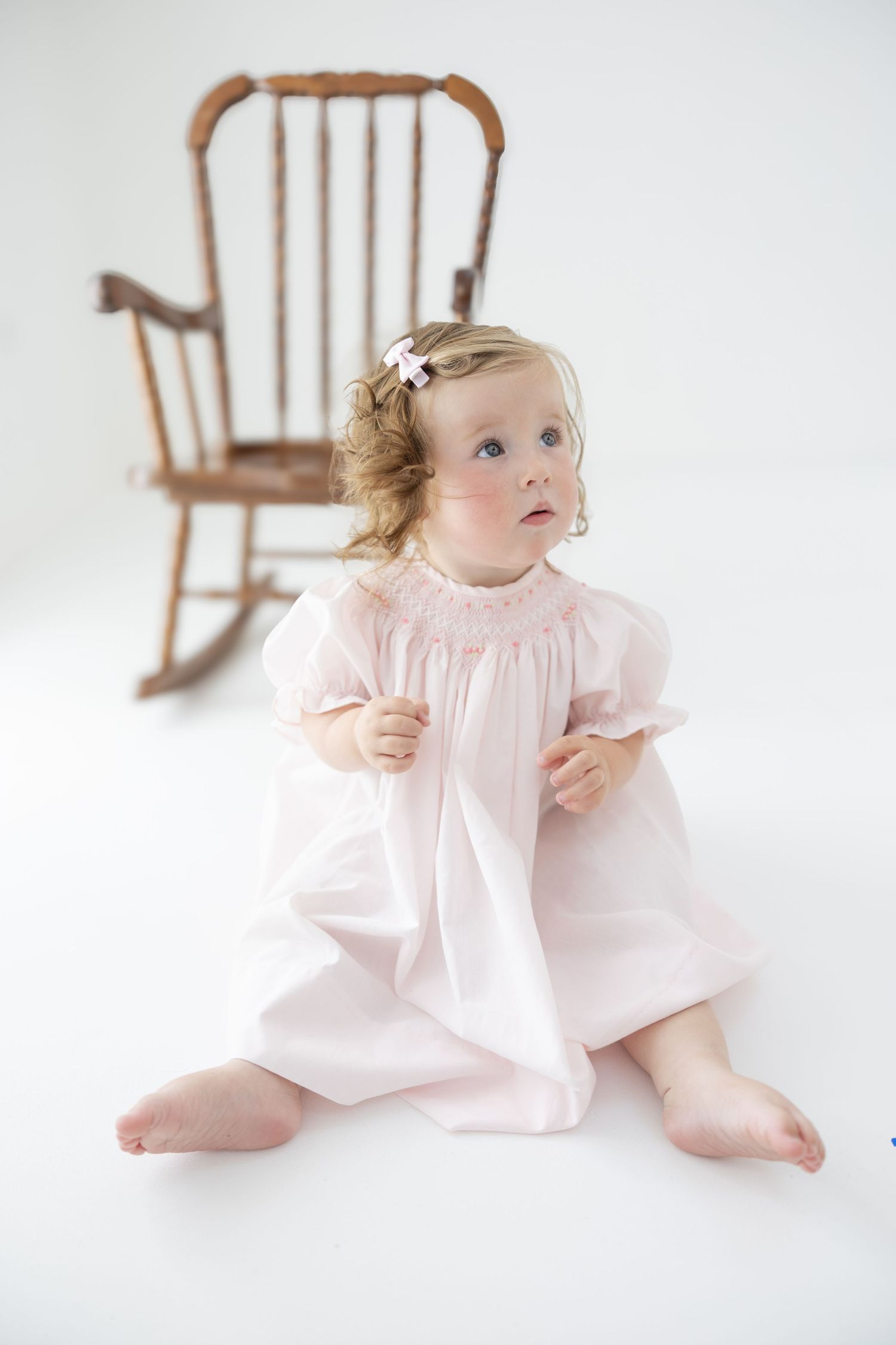 Toddler girl in pink smocked dress sitting barefoot on white floor with wooden rocking chair behind her.