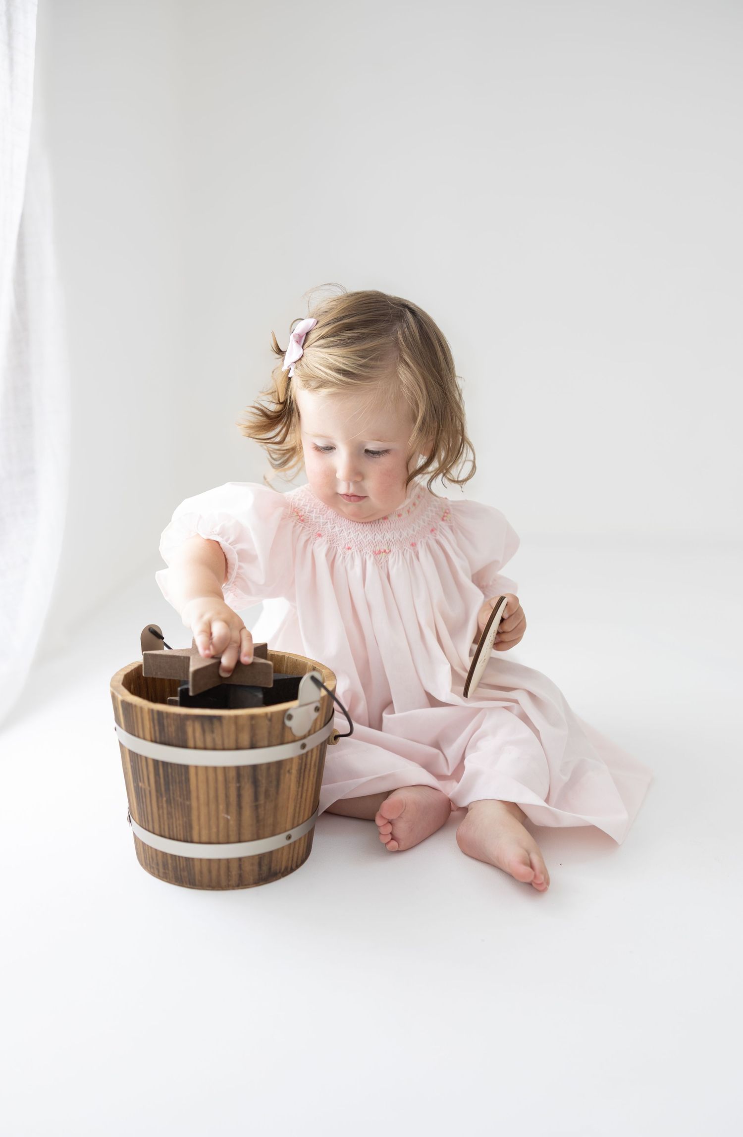 Toddler girl in pink smocked dress sitting on white floor, reaching into a rustic wooden bucket.