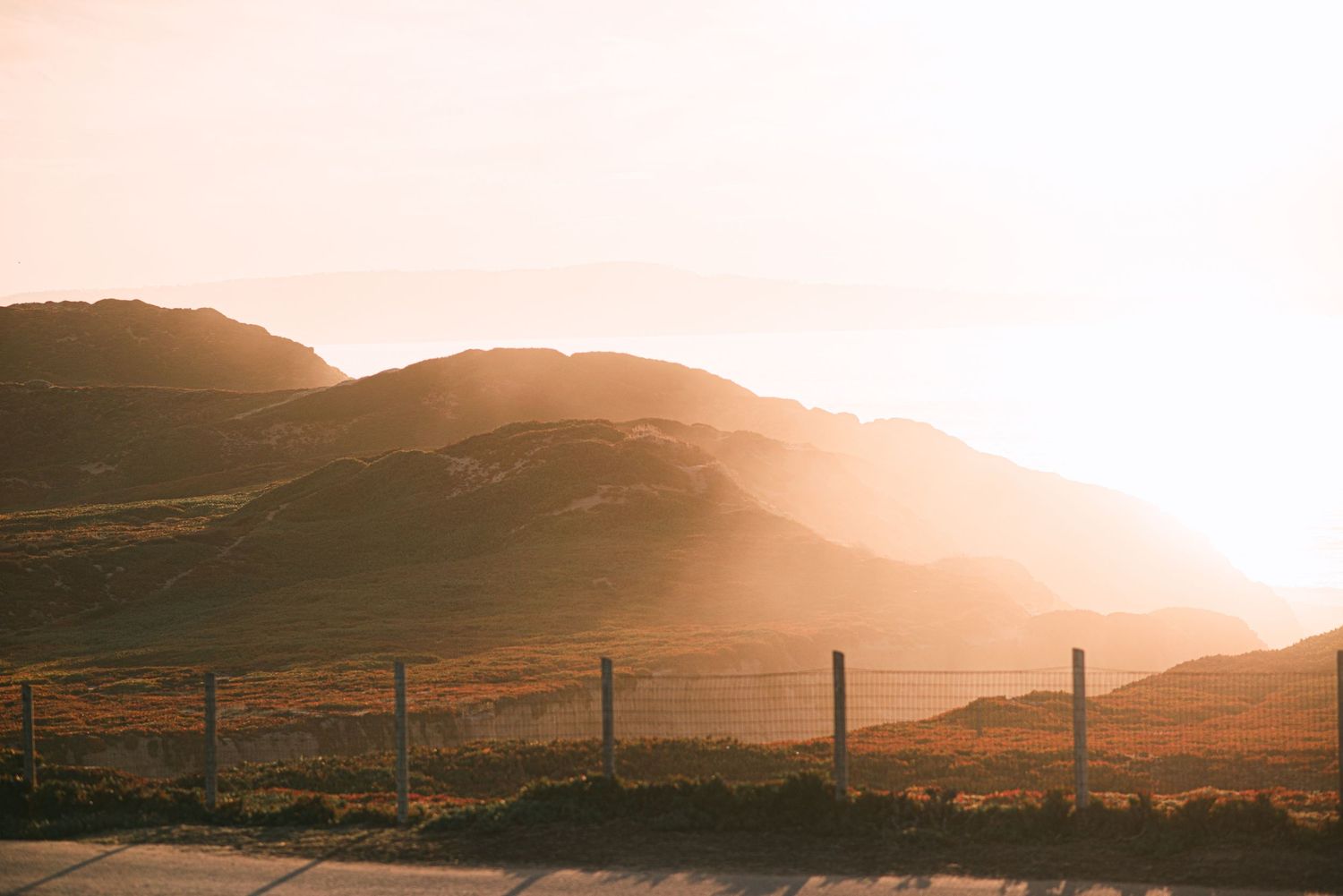 Golden sunset light illuminates rolling hills and a wire fence in a serene rural landscape.