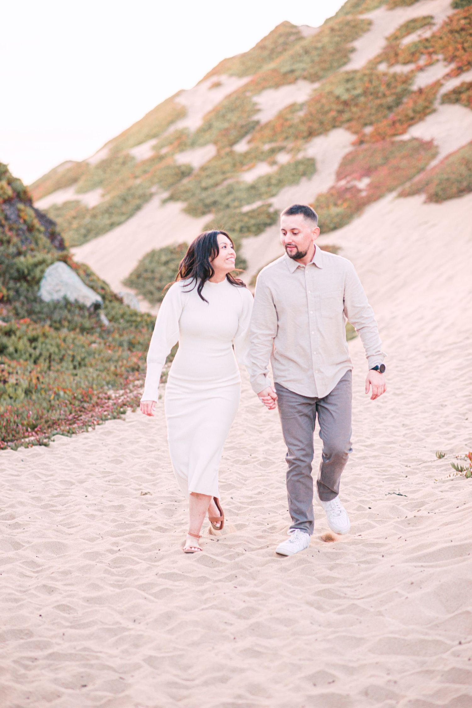 A couple walks hand in hand along a sandy coastal path during golden hour.