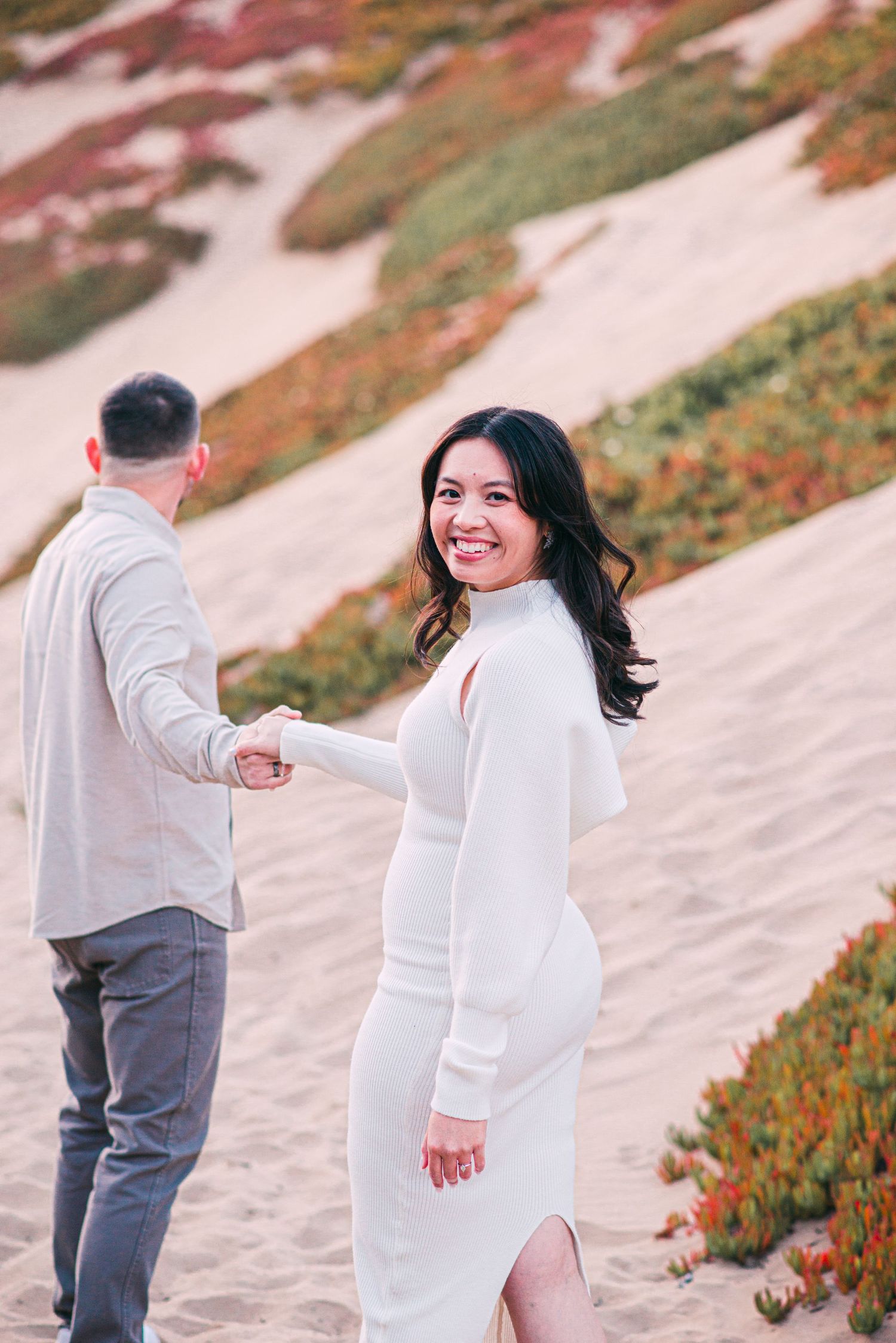Woman in white dress smiles while holding hands on coastal path with colorful vegetation.