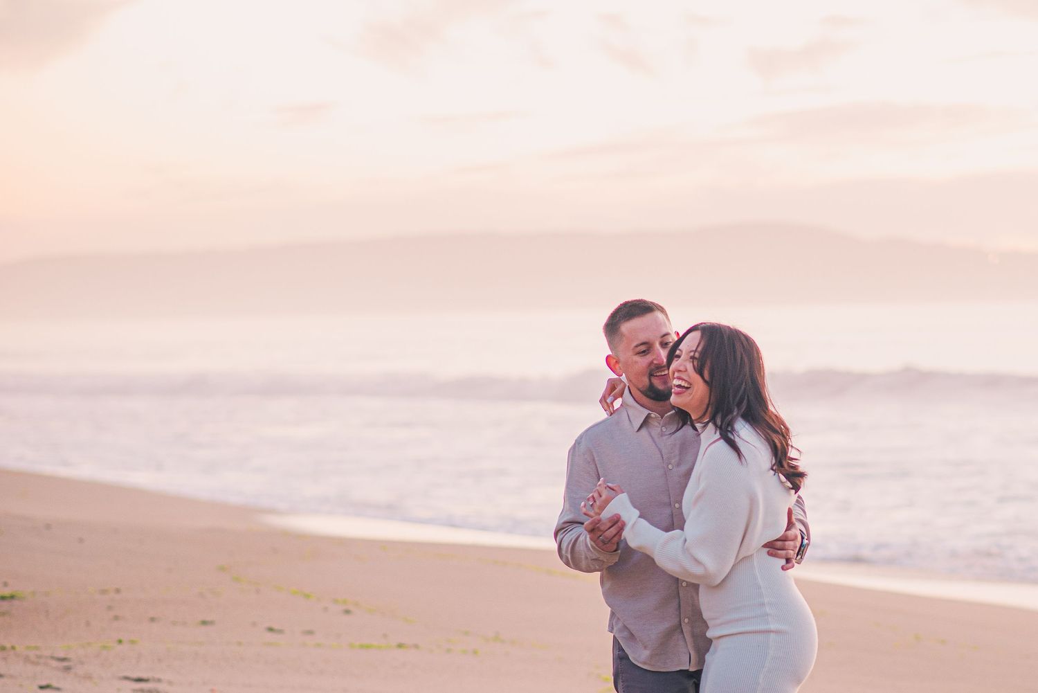 Romantic couple embracing on beach at sunset with soft pink sky and ocean waves in background.