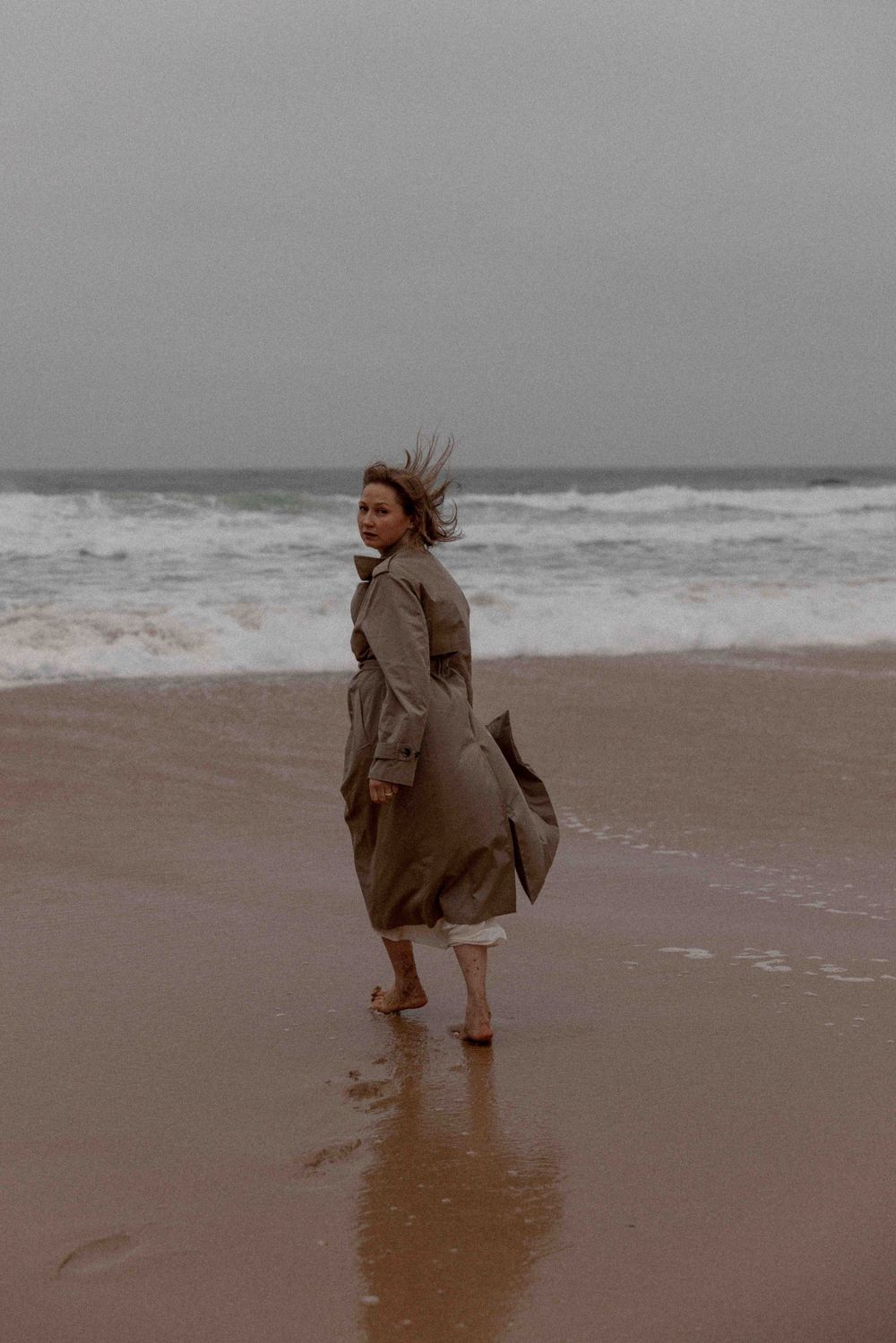 Person in flowing coat standing on windswept beach with waves crashing behind.