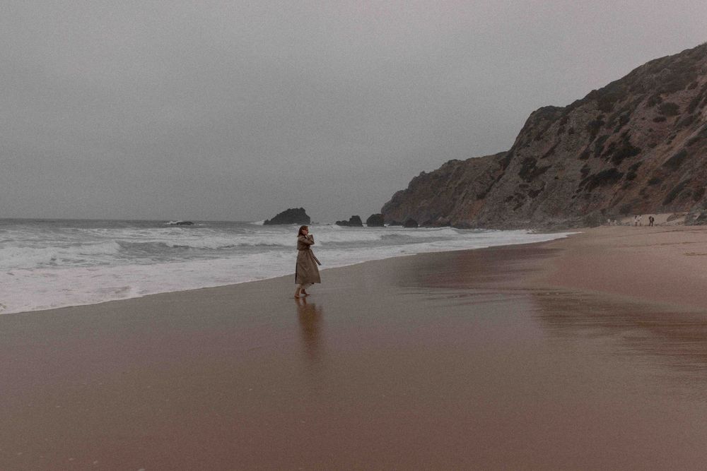 Figure standing alone on a misty beach with rocky cliffs in the background.