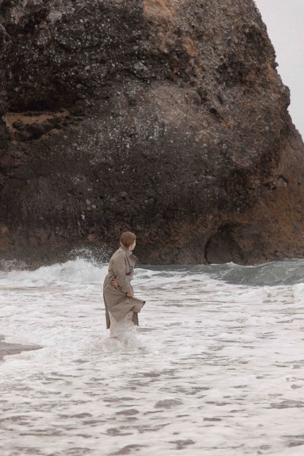 Figure wading in ocean waves near a large rocky cliff face.