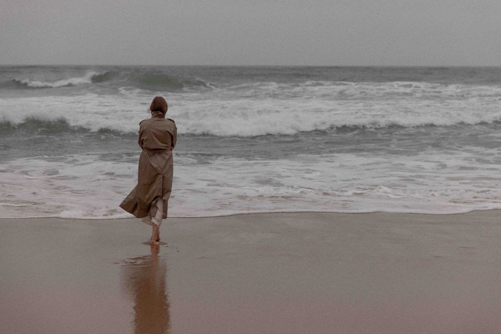 Figure in a flowing brown dress stands at the edge of crashing waves on a moody beach.