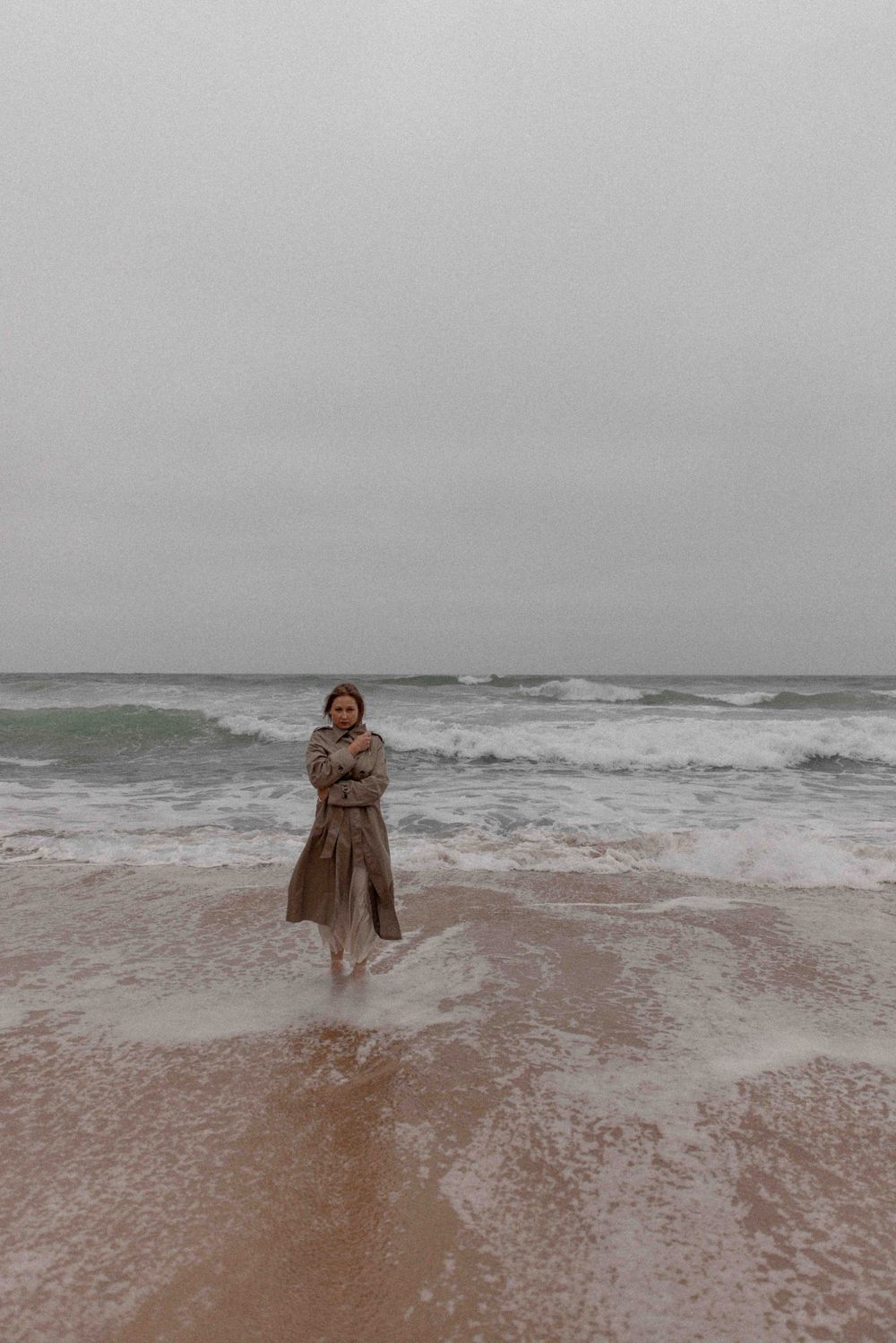 Person in a long coat stands ankle-deep in ocean surf on an overcast day.