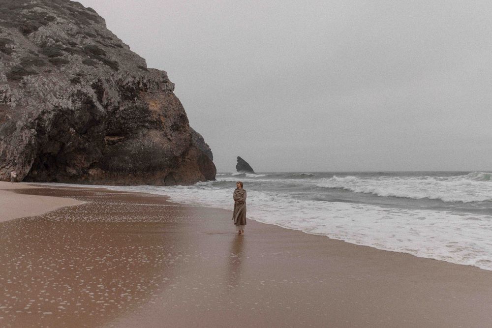 Solitary figure stands on beach near rocky cliffs in misty weather.